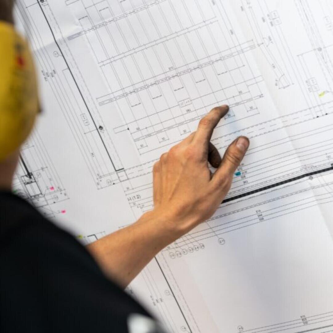 Eine Person, die einen gelben Gehörschutz trägt, zeigt mit einer leicht verschmutzten Hand auf einen Ausschnitt einer großen technischen Blaupause. Das Bild konzentriert sich auf die Hand und die Details der Blaupause.