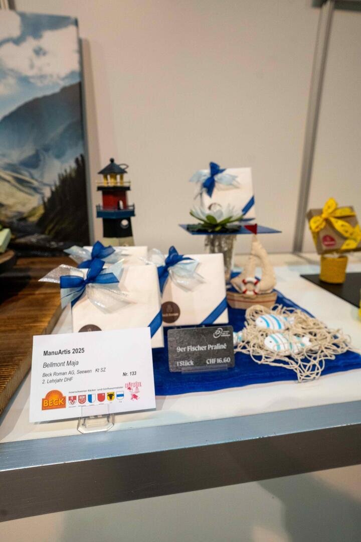 Auf einem Tisch, vor dem ein Etikett mit der Aufschrift „ManuArtis 2025“ steht, ist eine Auswahl an Geschenken mit maritimem Motiv ausgestellt, darunter Pralinen, ein Mini-Leuchtturm, Dekorationen aus Seil und ein kleiner Anker.