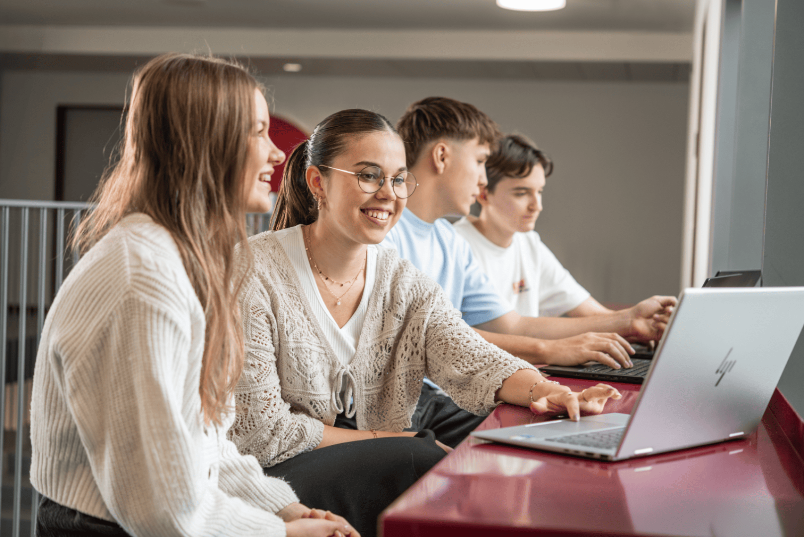 Vier junge Menschen sitzen an einem roten Tisch, arbeiten an Laptops und lächeln. Zwei sitzen im Vordergrund, beschäftigt und schauen auf einen Bildschirm, während zwei weitere hinter ihnen sitzen, ebenfalls konzentriert auf ihre Laptops.