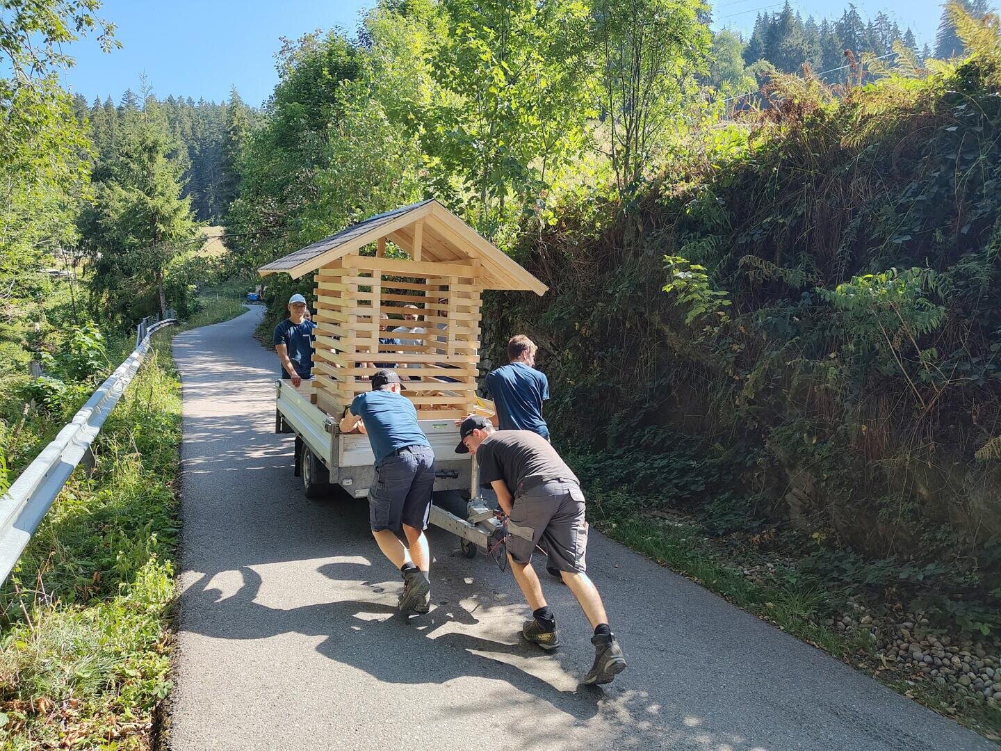 An einem sonnigen Tag schieben vier Personen eine Holzkonstruktion auf einem Anhänger einen gepflasterten Weg hinauf, der von Bäumen und Grünflächen gesäumt ist.