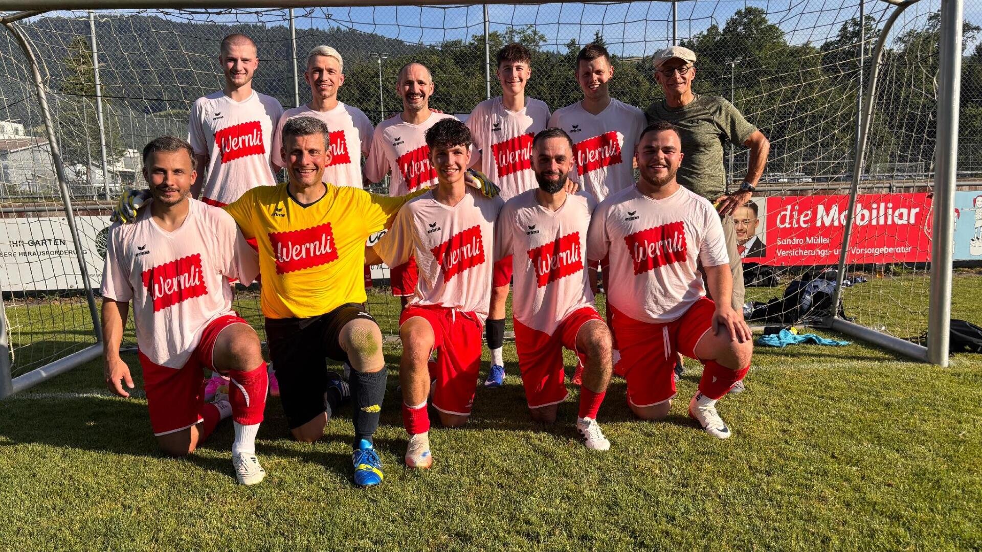 Eine Fußballmannschaft aus elf Männern posiert an einem sonnigen Tag vor einem Tor und trägt rot-weiße Trikots. Ein Spieler in Gelb sticht als Torwart hervor. Alle lächeln und knien oder stehen auf grünem Rasen.