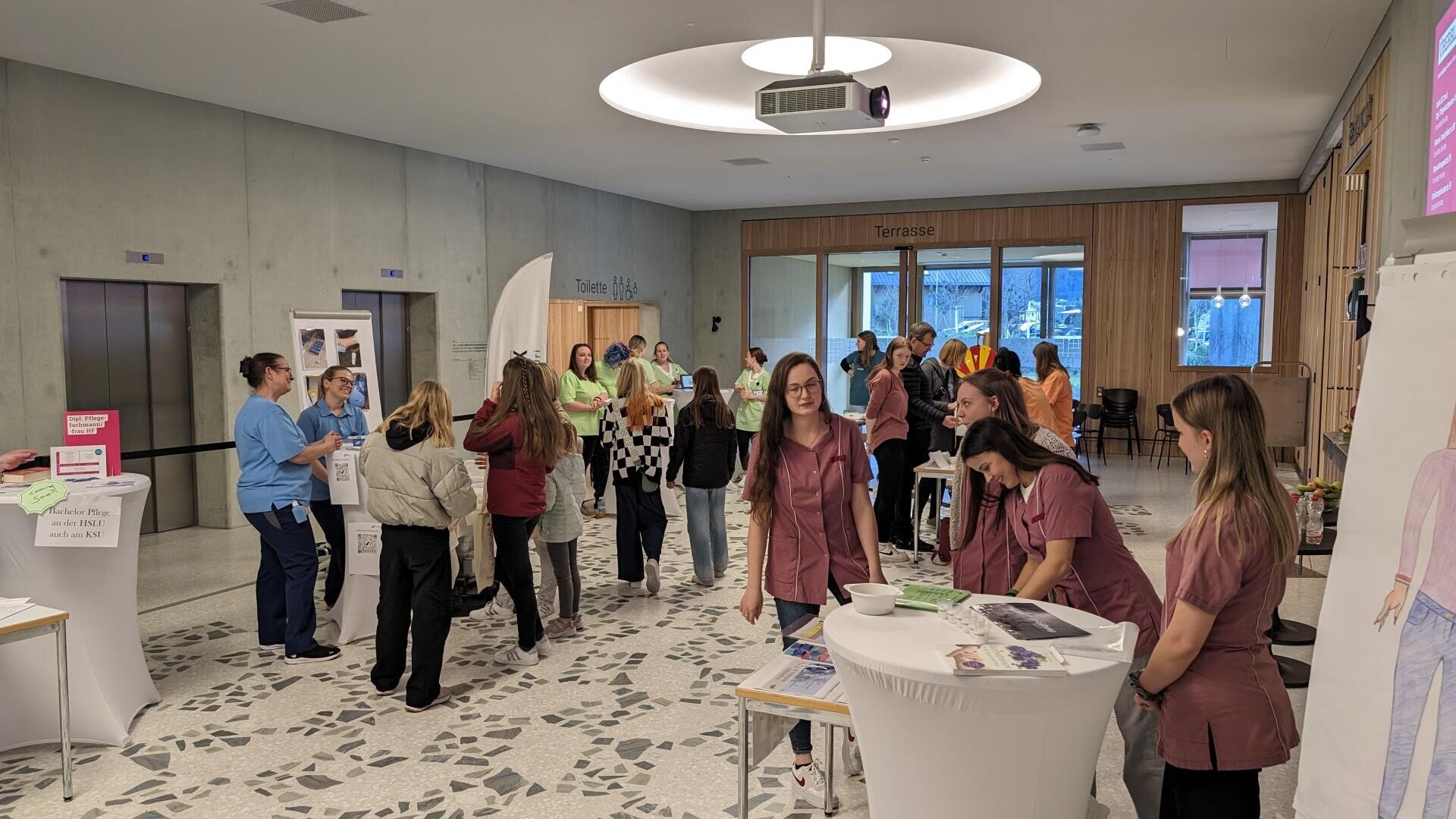 Eine geschäftige Veranstaltung in einer Halle mit Menschen, die sich an Ständen unterhalten. Mehrere Frauen in kastanienbraunen Uniformen stehen an einem runden Tisch im Vordergrund. Andere unterhalten sich und erkunden die Auslagen in einem modernen Raum mit hellen Wänden und Holzakzenten.