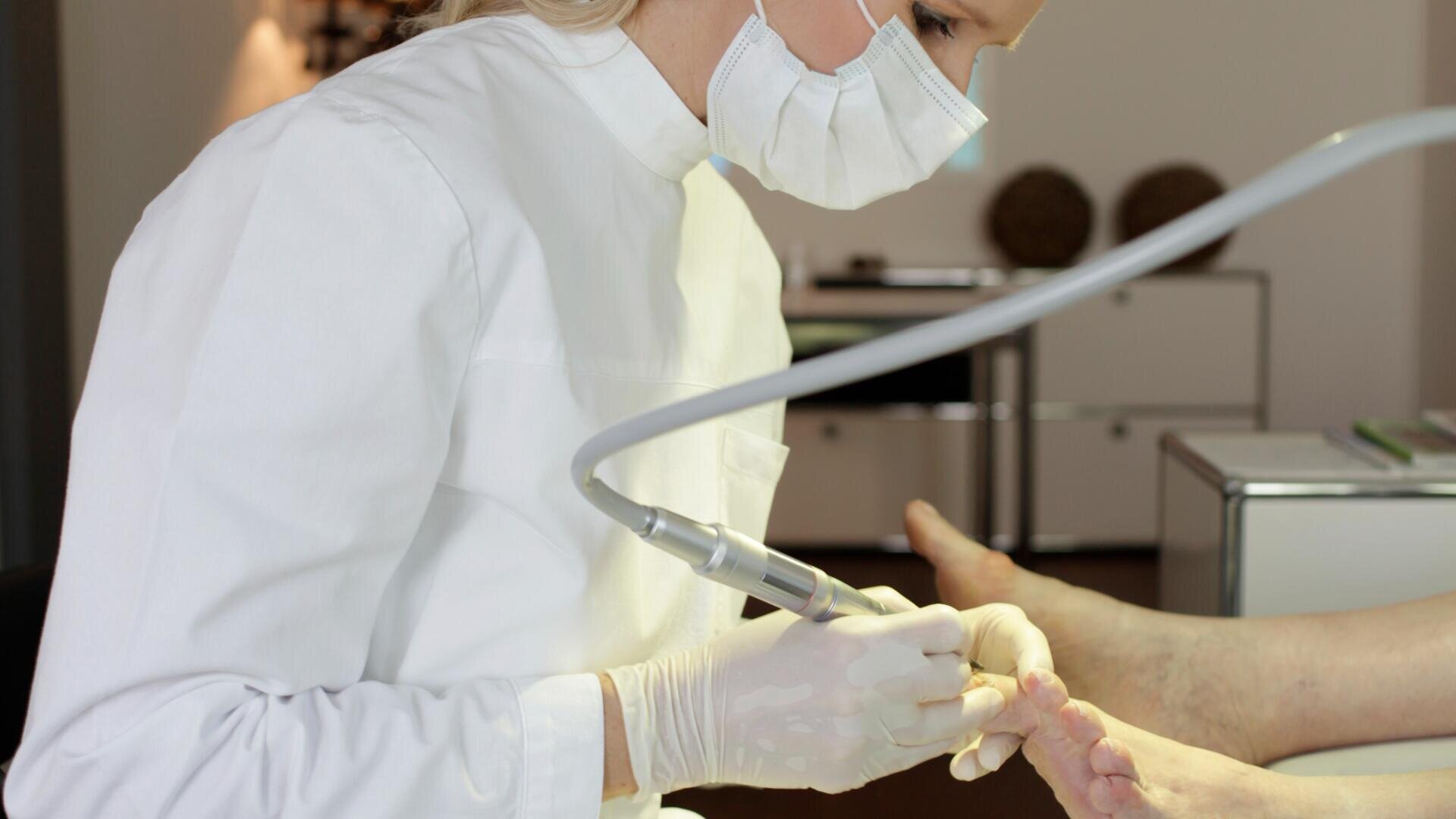 Eine Person, die eine Maske, Handschuhe und einen weißen Kittel trägt, behandelt in einer medizinischen Einrichtung mit einem zahnärztlichen oder medizinischen Instrument den nackten Fuß einer Person.