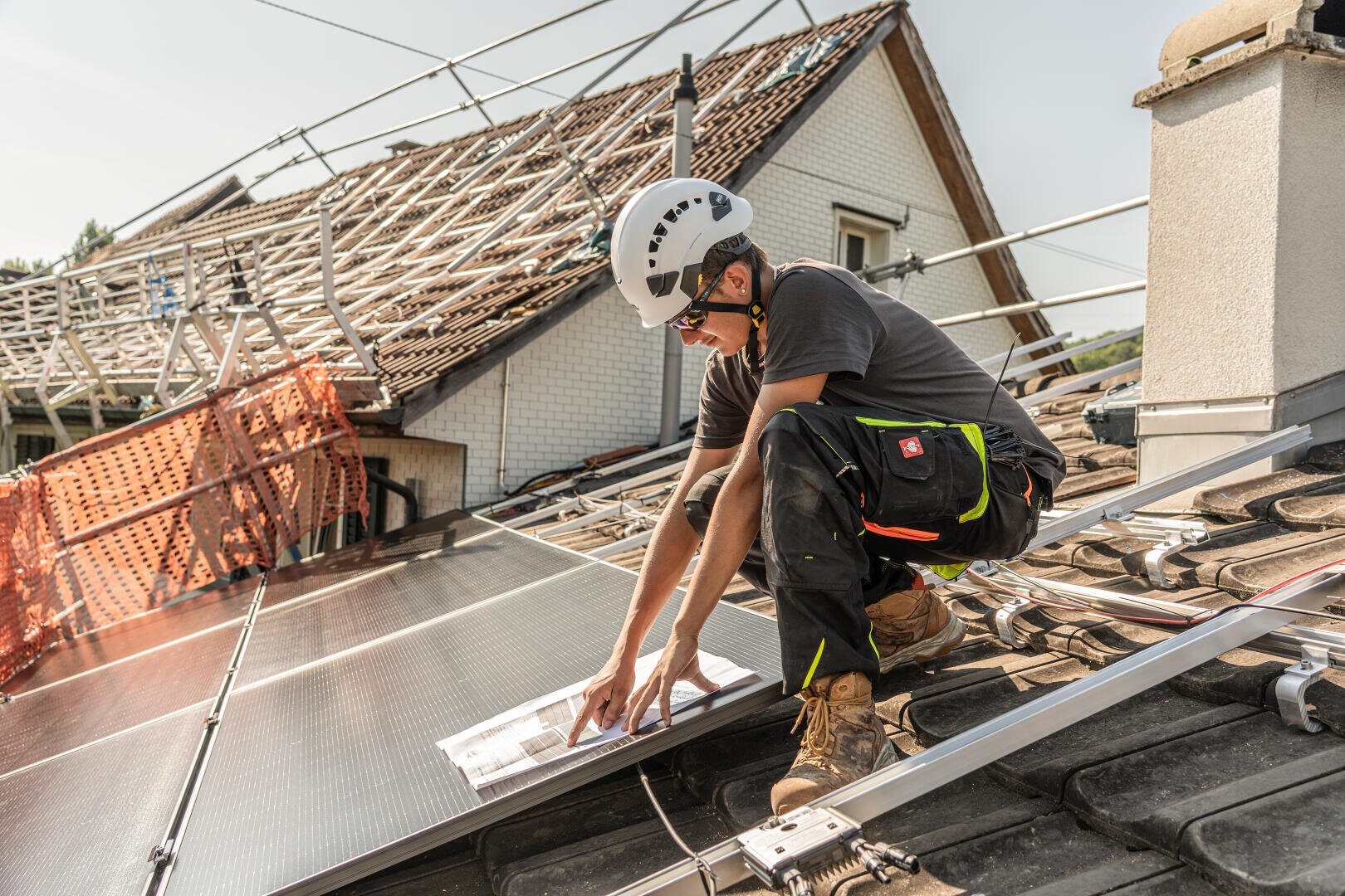 Ein Arbeiter mit Helm und Schutzausrüstung installiert Solarmodule auf einem schrägen Hausdach und orientiert sich dabei an einem Bauplan. Im Hintergrund ist ein weiteres Dach mit freiliegenden Dachsparren im Bau.