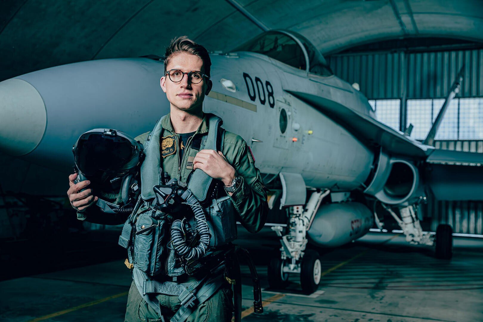 Ein Pilot im Fluganzug steht mit Helm und Ausrüstung in einem Hangar, hinter ihm ist ein Kampfjet mit der Nummer 008 geparkt. Licht scheint durch die Fenster des Hangars und erhellt die Szene.