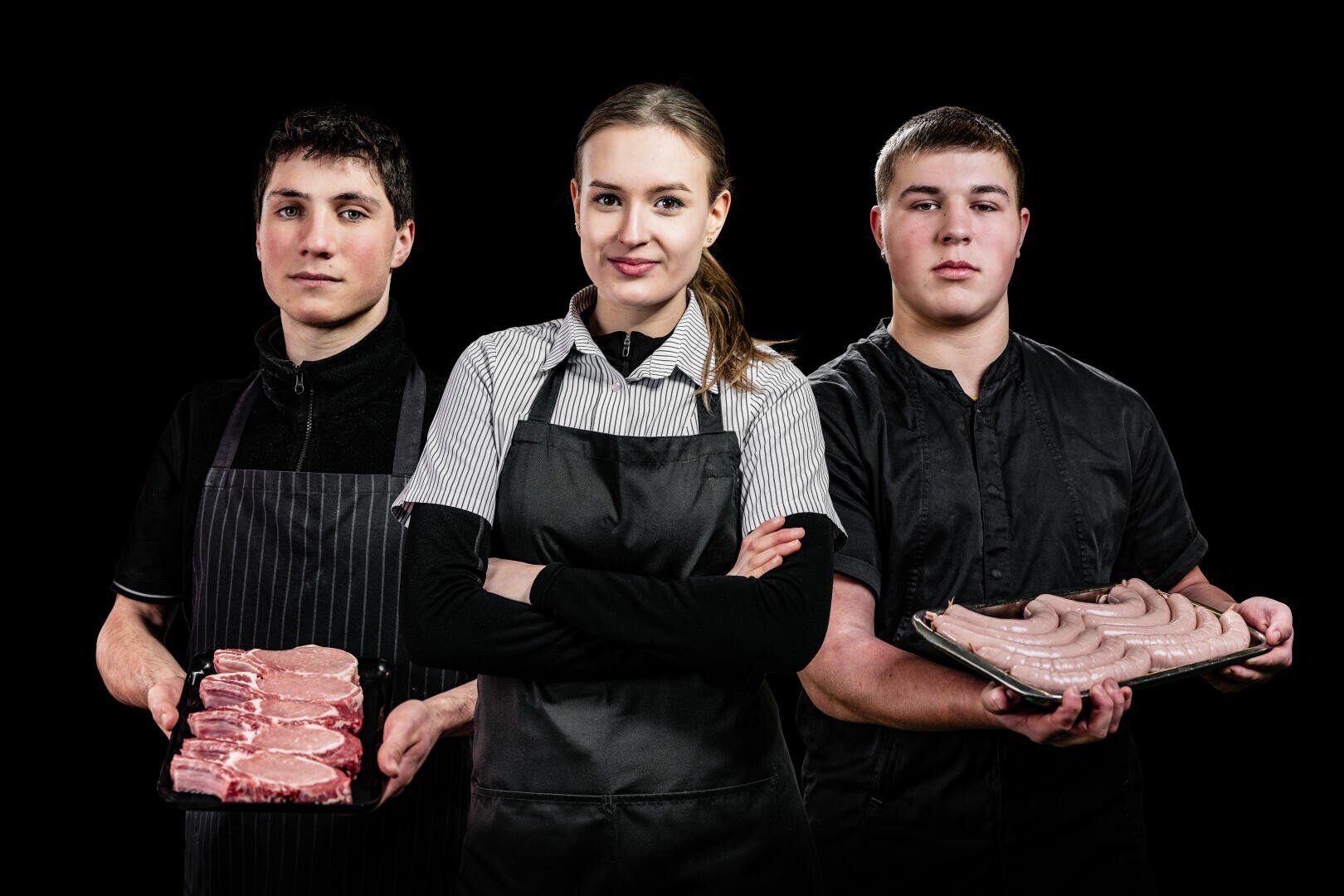Drei Metzger in schwarzen Uniformen stehen zusammen vor einem schwarzen Hintergrund. Die Person in der Mitte steht mit verschränkten Armen, während die beiden anderen Tabletts mit rohem Fleisch und Würsten halten.