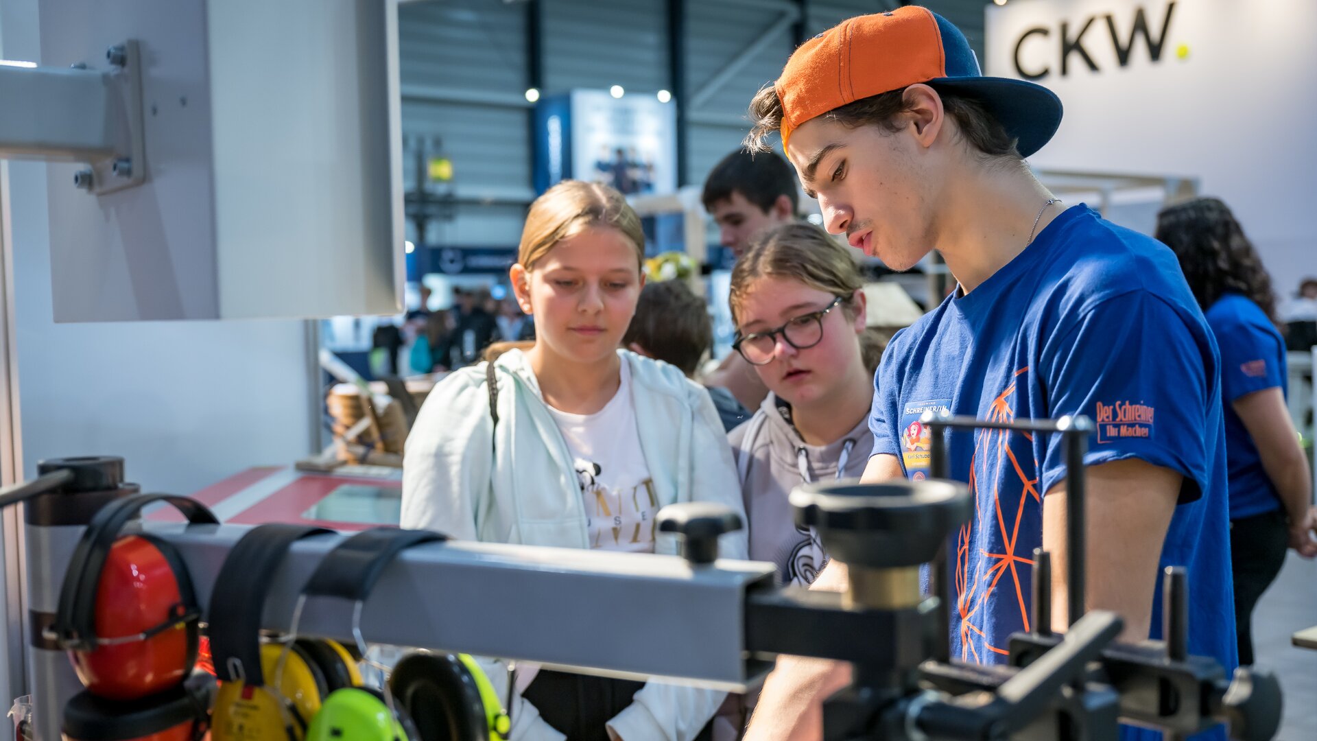 Ein junger Mann mit blauem Hemd und orangefarbener Mütze erklärt zwei interessierten Mädchen an einem Stand in einer hell erleuchteten Halle oder auf einer Messe die Geräte, während im Hintergrund weitere Personen zu sehen sind.