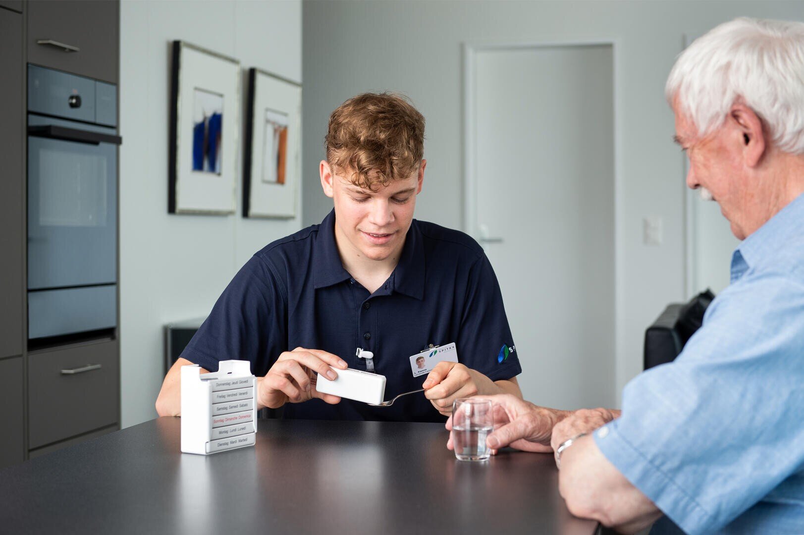 Eine junge Pflegekraft hilft einem älteren Mann bei der Einnahme von Medikamenten und zeigt ihm einen Tabletten-Organizer auf einem Tisch. Der ältere Mann hält ein Glas Wasser in der Hand. Sie befinden sich in einem modernen, hellen Raum.