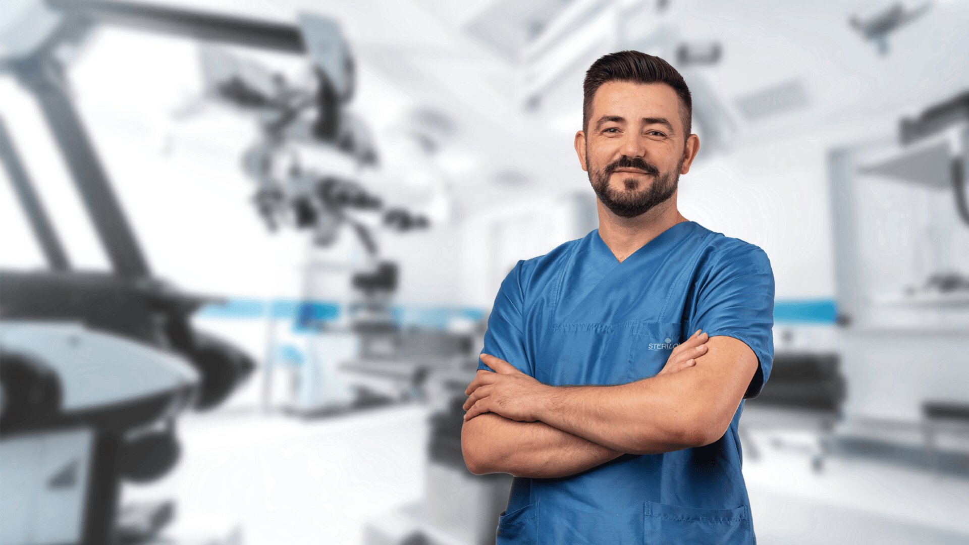 Eine selbstbewusste männliche medizinische Fachkraft in blauem Kittel steht mit verschränkten Armen lächelnd in einer hellen, modernen medizinischen Einrichtung mit fortschrittlichen Geräten im Hintergrund.