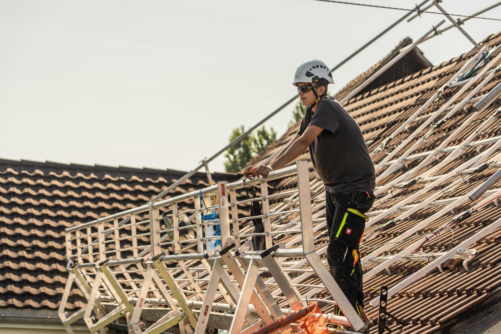 Ein Arbeiter mit Helm und Schutzausrüstung steht auf einer Hebebühne und arbeitet auf dem Schrägdach eines Hauses, das gerade renoviert wird, wobei die Dachziegel entfernt und ein Gerüst aufgestellt wurde.