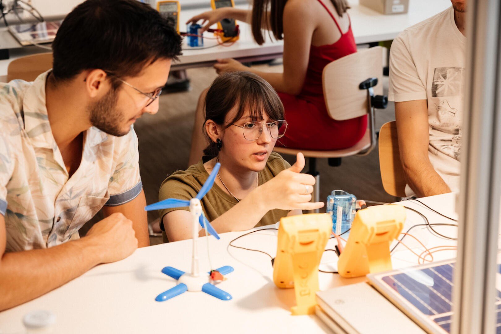 Ein junges Mädchen zeigt den Daumen nach oben, während es mit zwei jungen Erwachsenen an einem Tisch an einem wissenschaftlichen Projekt arbeitet, umgeben von Lehrmitteln wie einer Mini-Windturbine, Messgeräten und einem Solarpanel in einem Klassenzimmer.