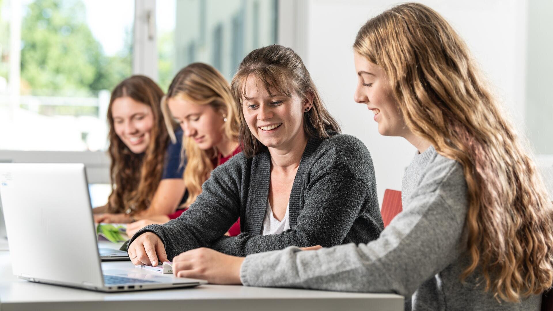 Vier Frauen sitzen an einem Tisch, lächeln und arbeiten zusammen. Zwei konzentrieren sich auf einen Laptop im Vordergrund, während die beiden anderen im Hintergrund in Notizbücher schreiben. Die Umgebung wirkt hell und modern.