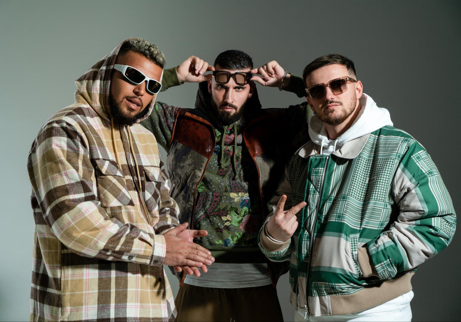 Drei Männer in stylischen Streetwear-Jacken und mit Sonnenbrillen posieren selbstbewusst vor einem schlichten Hintergrund. Der Mann in der Mitte rückt seine Brille zurecht, während die anderen mit den Händen gestikulieren.
