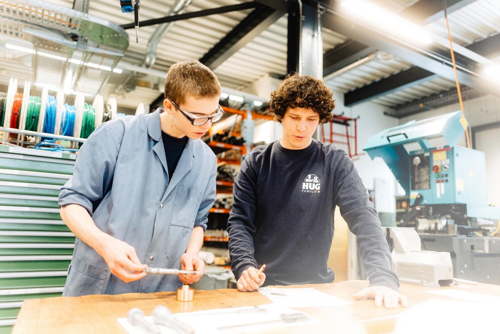 Zwei junge Männer arbeiten gemeinsam an einem Tisch in einer Werkstatt. Einer trägt eine Schutzbrille und einen Laborkittel und misst ein Teil, während der andere auf Papiere auf dem Tisch zeigt. Im Hintergrund sind Werkzeuge und Maschinen zu sehen.