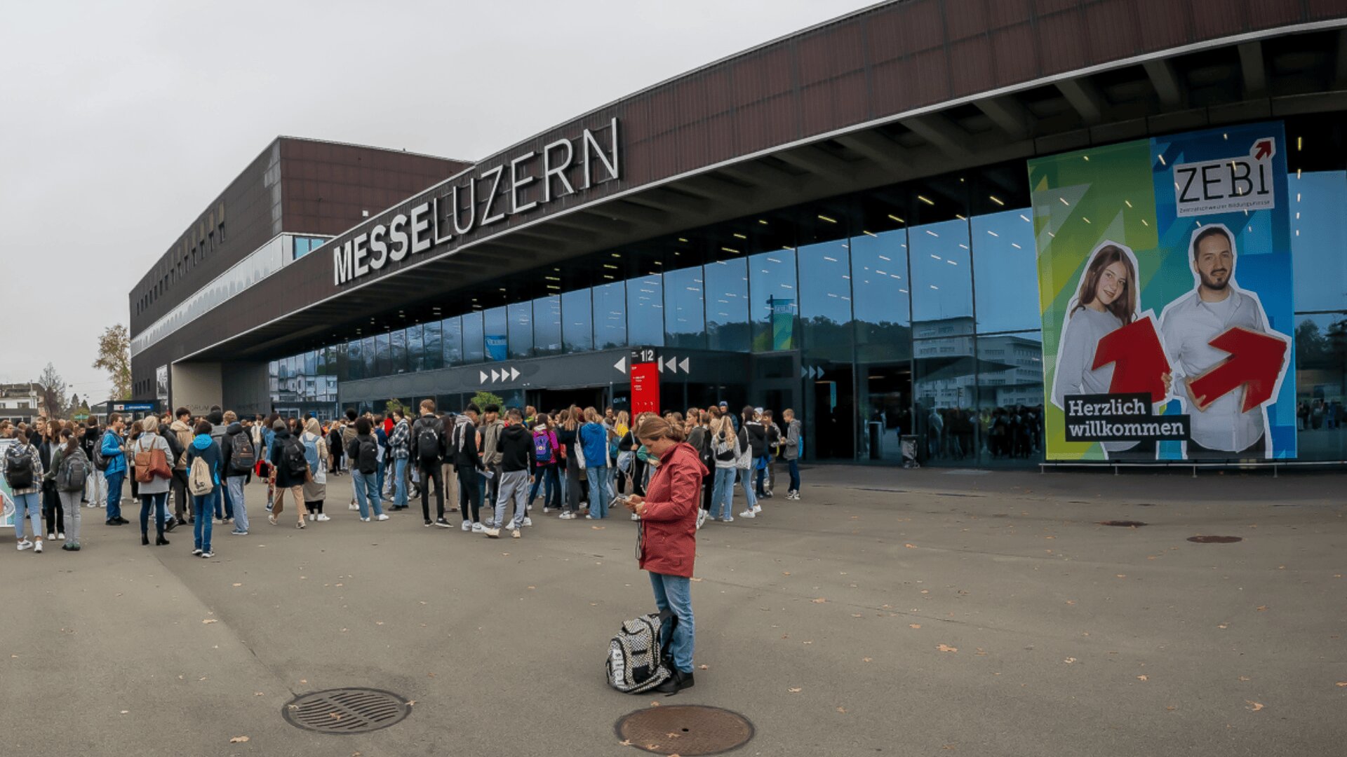 Eine Menschenmenge versammelt sich vor der Messehalle Luzern. In den großen Glasfenstern spiegelt sich die Umgebung. Auf einem Transparent mit zwei lächelnden Menschen und roten Pfeilen steht "Herzlich willkommen". Eine Person steht davor und bedient ein Telefon.