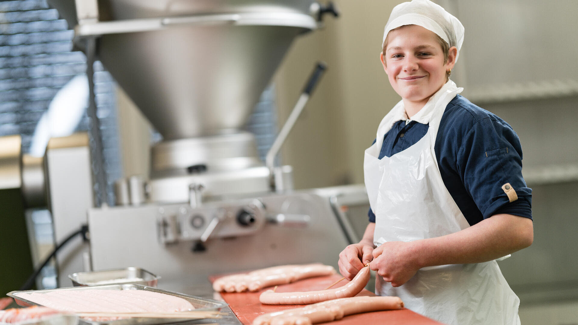 Eine lächelnde Arbeiterin mit weißer Schürze und Haarnetz formt in einem Lebensmittelverarbeitungsbetrieb Würste von Hand, während auf dem Tisch Wurstherstellungsgeräte und Därme zu sehen sind.