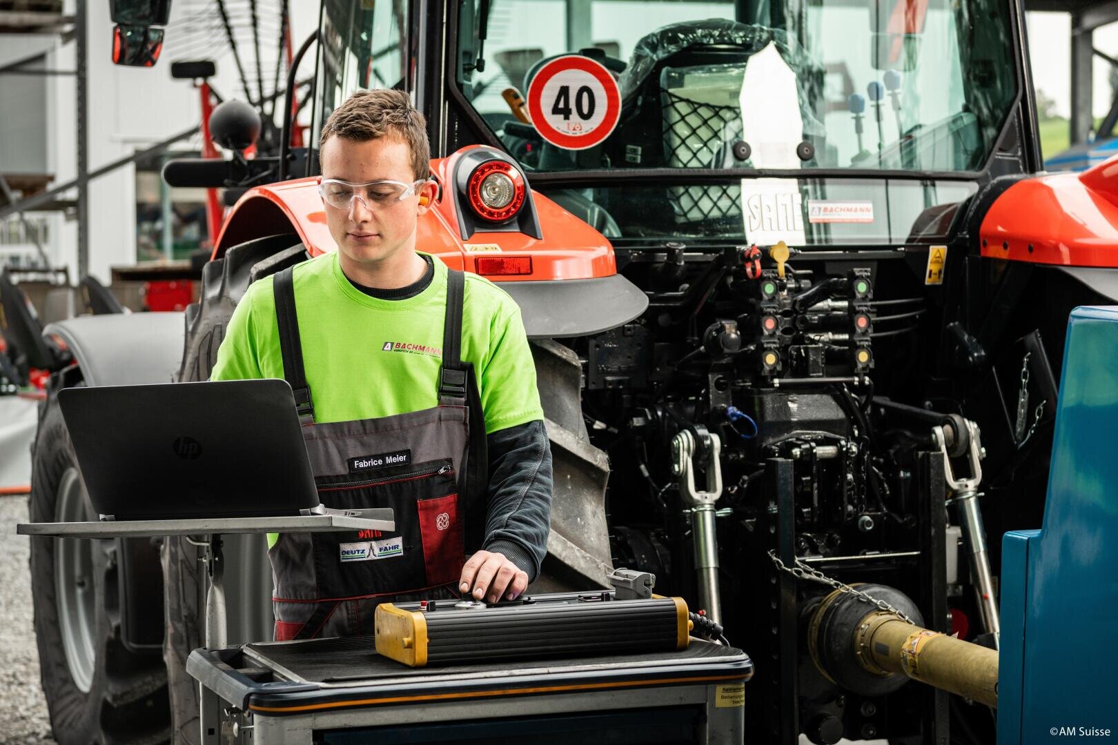 Ein junger Mechaniker mit Schutzbrille und Arbeitsoverall arbeitet mit einem Laptop neben einem roten Traktor und führt Diagnose- oder Wartungsarbeiten in einer Werkstatt im Freien durch.
