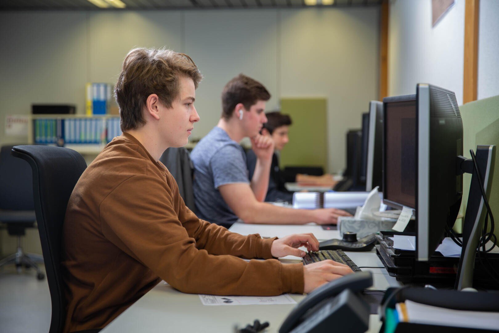 Drei junge Männer sitzen an Schreibtischen in einem Büro und arbeiten an Desktop-Computern. Einer trägt einen braunen Pullover und benutzt eine Maus, während ein anderer Ohrstöpsel trägt und auf seinen Bildschirm konzentriert ist. Auf den Schreibtischen liegen Büromaterialien.