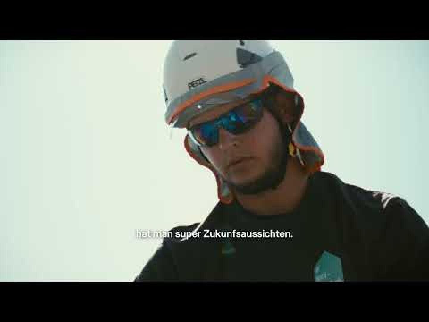 Eine Person, die einen Helm, eine Sonnenbrille und ein dunkles Hemd trägt, schaut nach unten. Die Bildunterschrift lautet: „Der Mann mit dem Hut – super Zukunftsaussichten“. Der Hintergrund ist schlicht und hell beleuchtet.