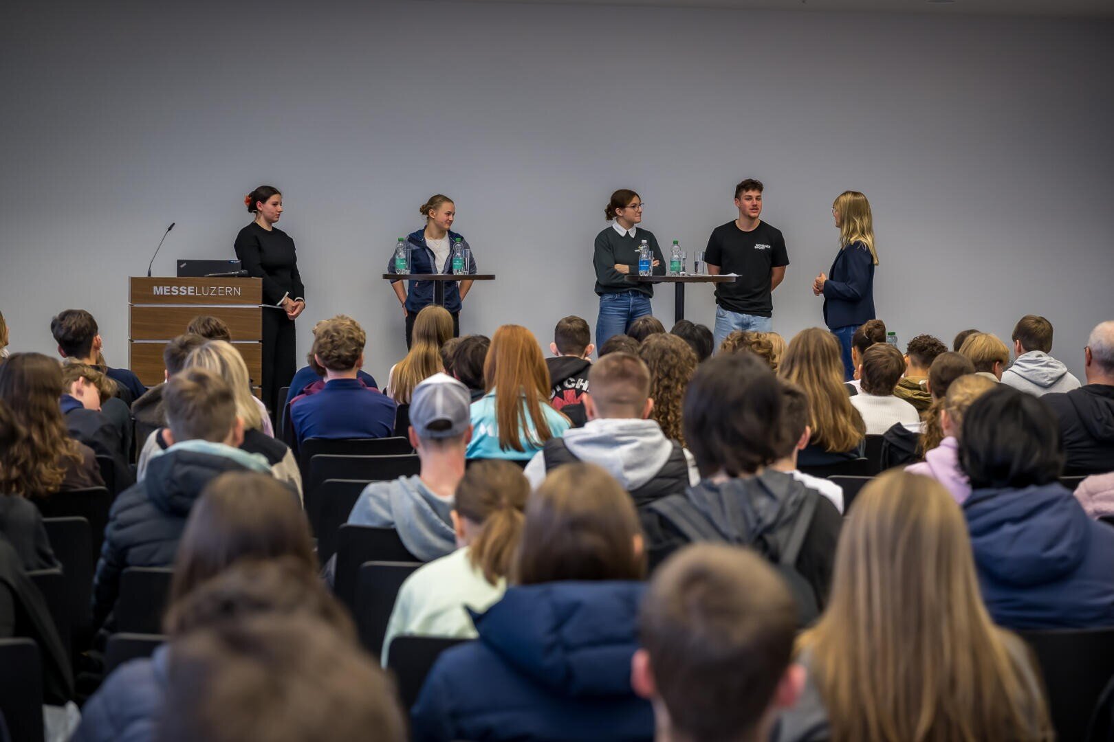 Eine Gruppe von fünf Personen steht und spricht auf einer Bühne vor einem Publikum in einem Konferenzraum. Eine Person steht an einem Podium mit der Aufschrift Messe Luzern, während das Publikum zuschaut.
