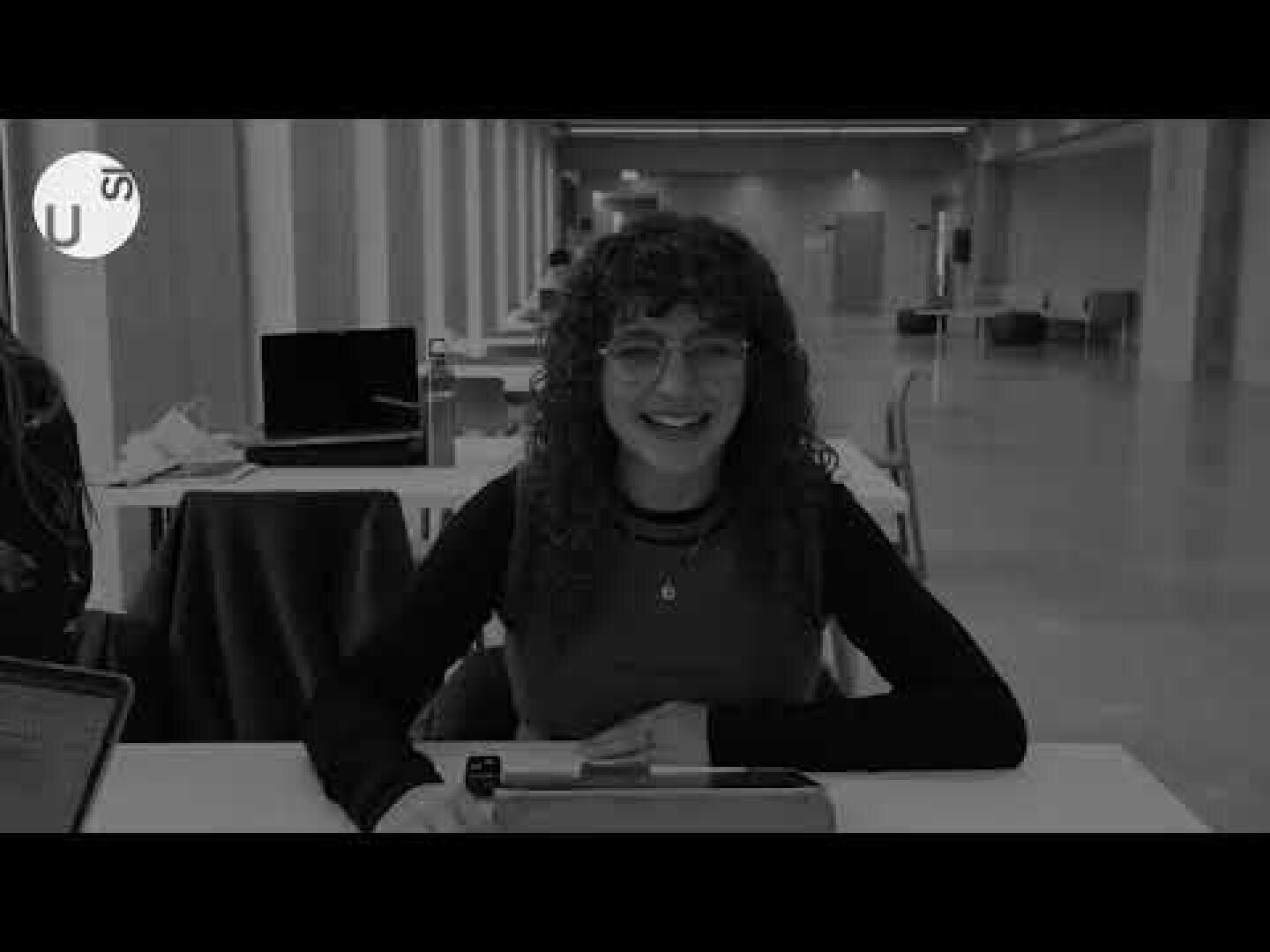 Eine Person mit lockigem Haar und Brille lächelt, während sie an einem Schreibtisch in einem geräumigen, modernen Raum sitzt, in dem im Hintergrund Laptops und Stühle zu sehen sind.