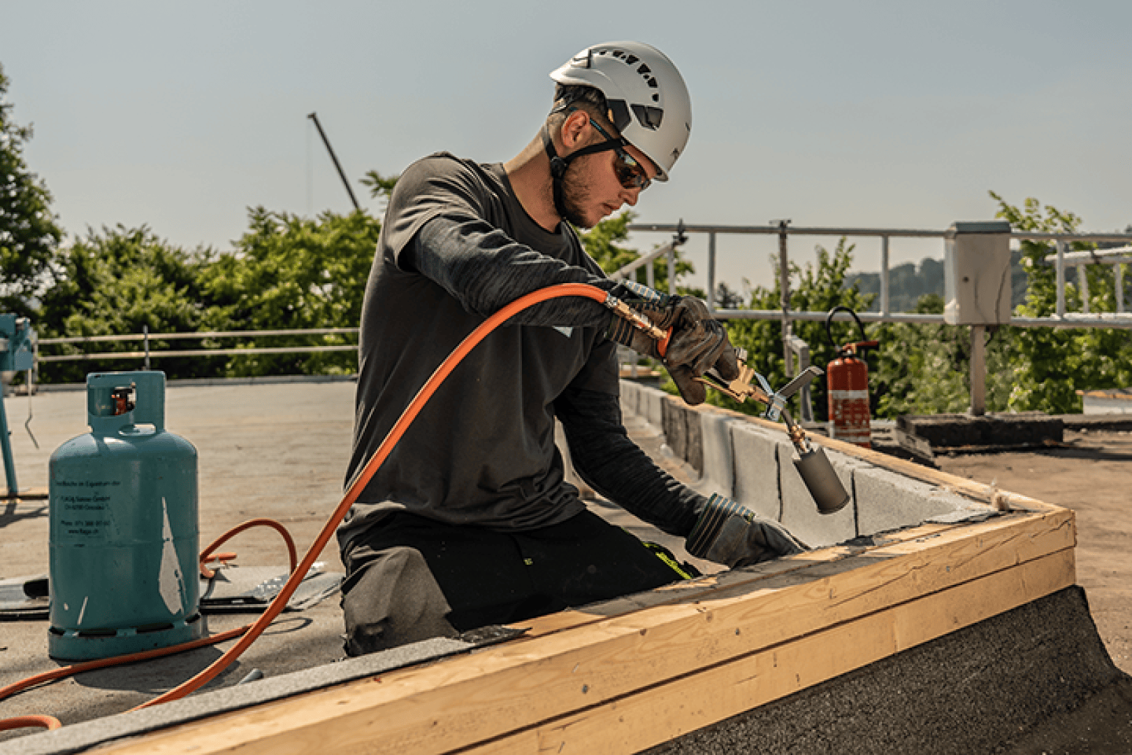 Ein Bauarbeiter, der einen Helm und eine Sonnenbrille trägt, benutzt einen an eine Gasflasche angeschlossenen Brenner, während er bei sonnigem Wetter auf einem Dach arbeitet. Im Hintergrund sind Bäume und Sicherheitsgeländer zu sehen.