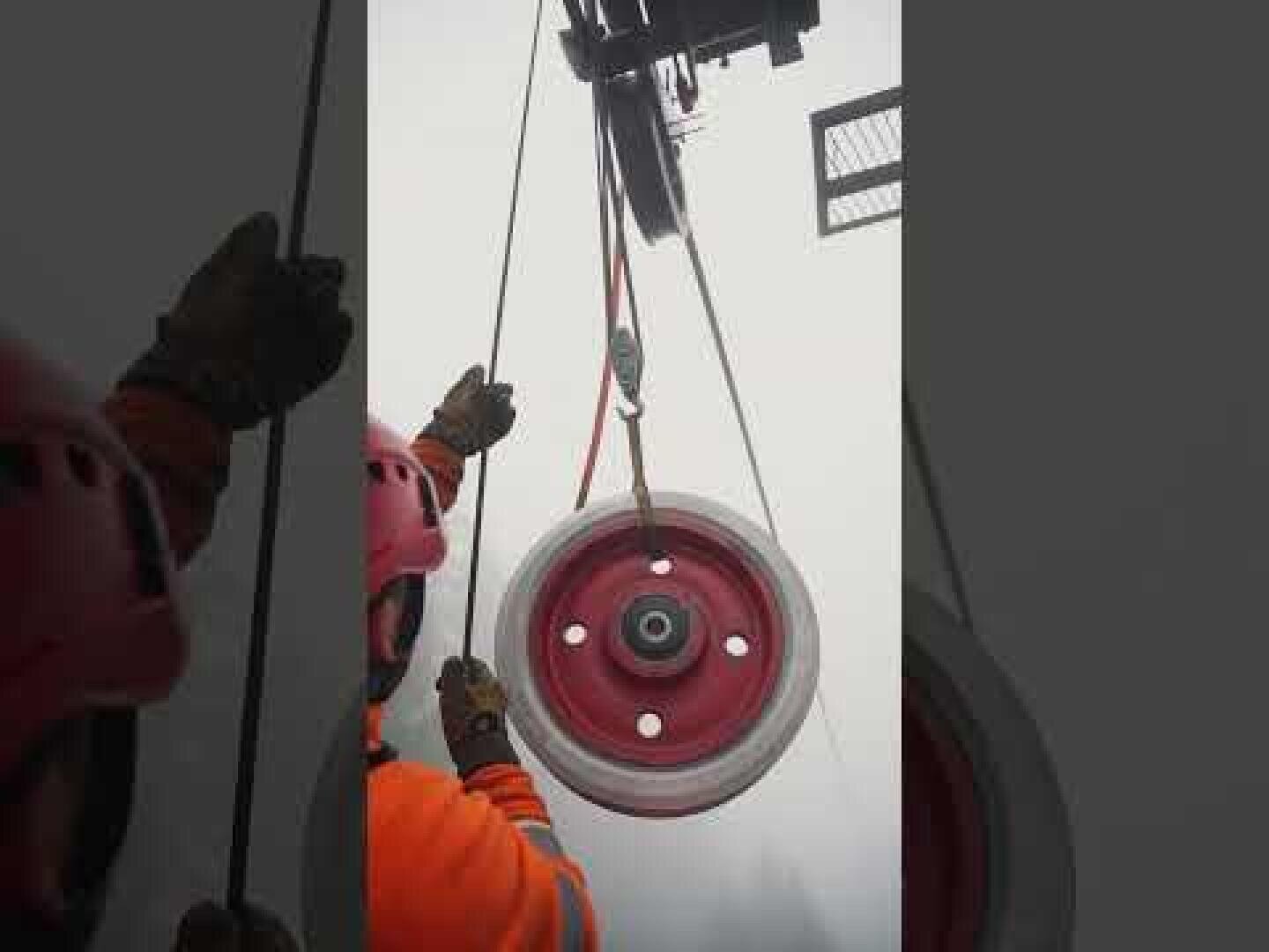 Zwei Arbeiter in orangefarbener Schutzkleidung und mit Helmen führen mithilfe von Seilen eine große rote Seilrolle, die von einem Kran vor einem nebligen oder bewölkten Himmel angehoben wird.