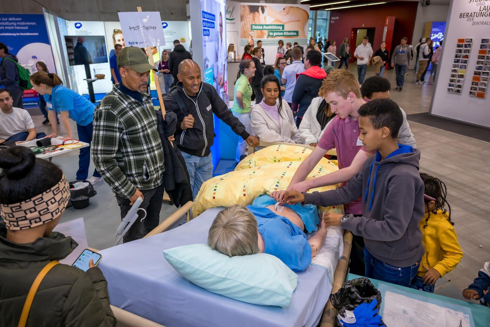 Eine Gruppe von Kindern und Erwachsenen versammelt sich um ein Krankenhausbett mit einer Patientenpuppe und nimmt an einer praktischen Bildungsaktivität bei einer belebten Indoor-Veranstaltung teil. Im Hintergrund sind Informationsstände und Menschen zu sehen.
