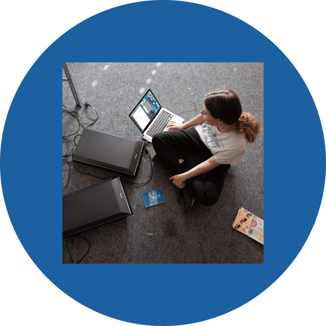 Eine Person sitzt auf einem Teppichboden mit einem Laptop, umgeben von zwei großen Scannern, einer blauen Karte und einem aufgeschlagenen Magazin, alles in einem runden blauen Rahmen.
