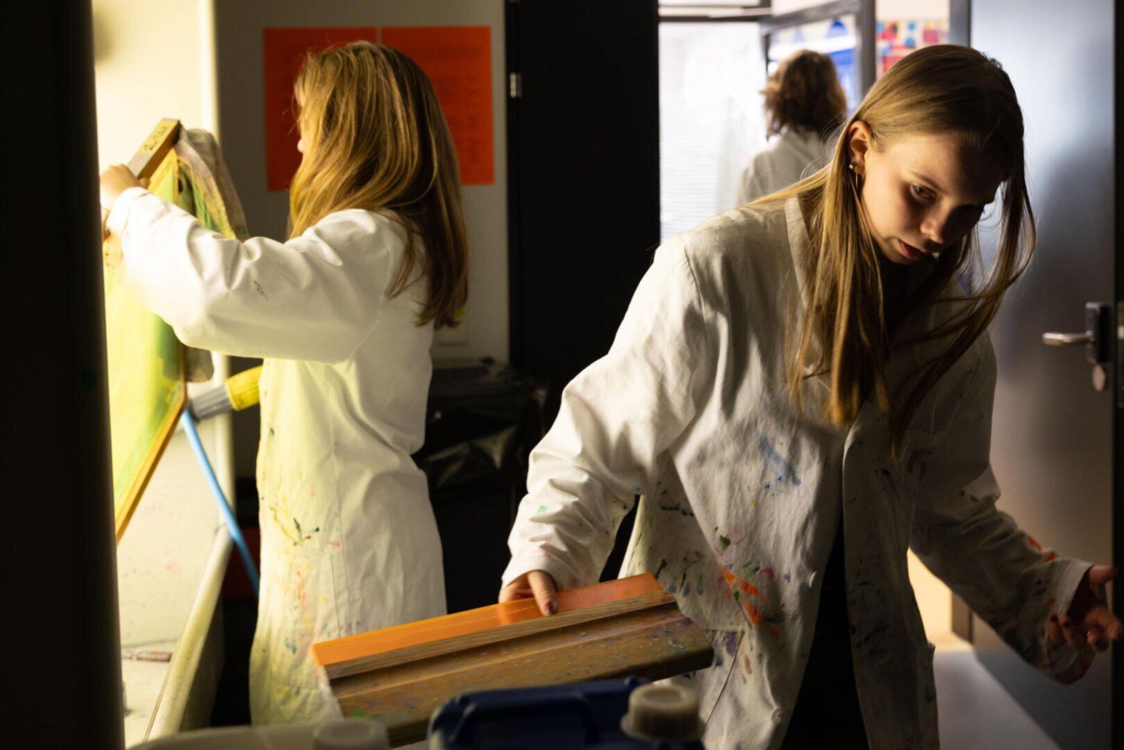 Zwei Personen in weißen Laborkitteln arbeiten in einem gut beleuchteten Raum an Kunstprojekten. Die eine hält einen Siebdruckrahmen über einen Tisch, während die andere einen weiteren Rahmen vorbereitet, während im Hintergrund Kunstzubehör und eine dritte Person zu sehen sind.