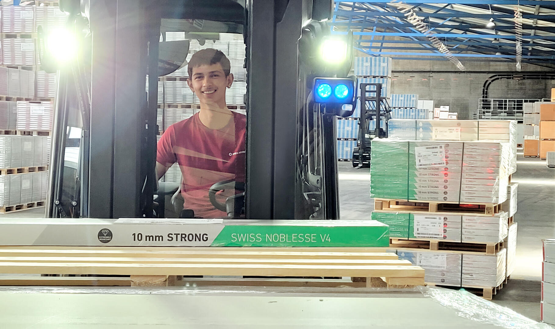 Eine lächelnde Person in einem roten Hemd bedient einen Gabelstapler in einem Lagerhaus und bewegt eine Palette mit Swiss Noblesse V4 10mm starken Platten. Im Hintergrund sind Stapel von Kisten und Paletten zu sehen.