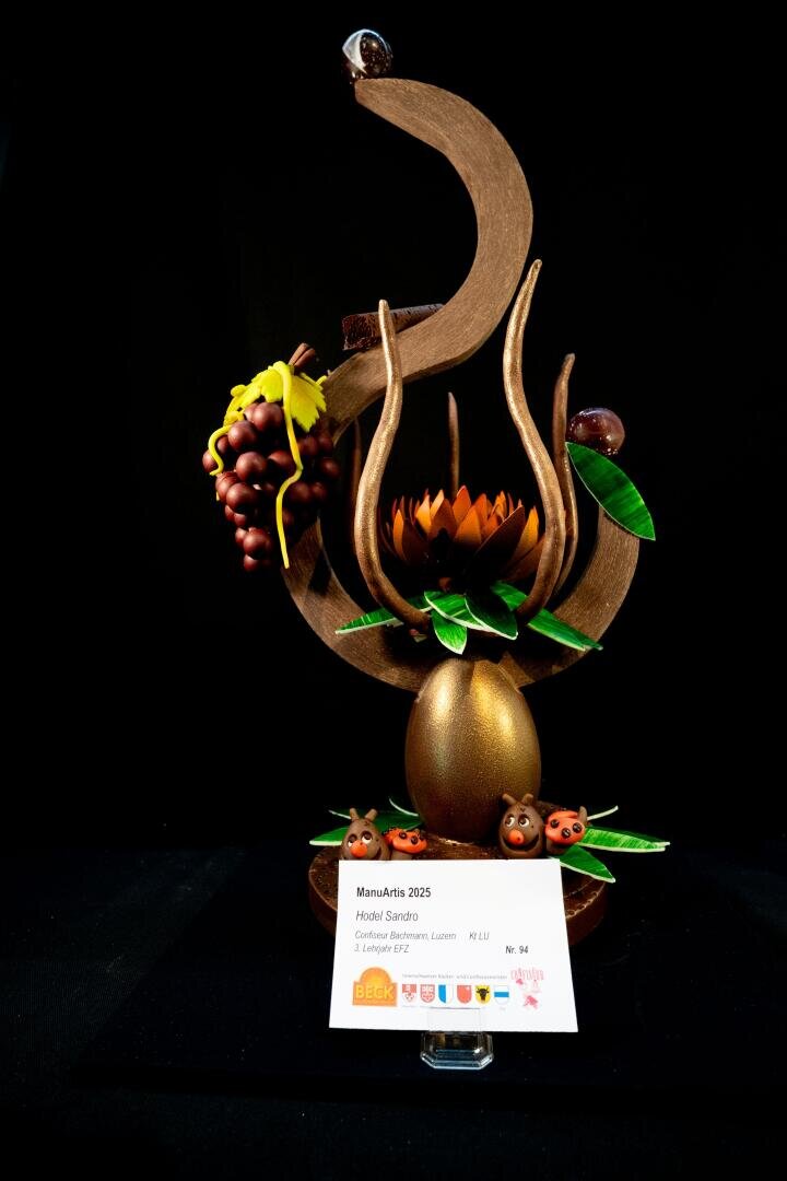 Eine Schokoladenskulptur mit geschwungenen Formen, ein goldenes Ei, eine Blume, Marienkäfer, Weintrauben, grüne Blätter und eine Karte mit der Aufschrift „MamuArtis 2025“. Der Hintergrund ist schwarz.
