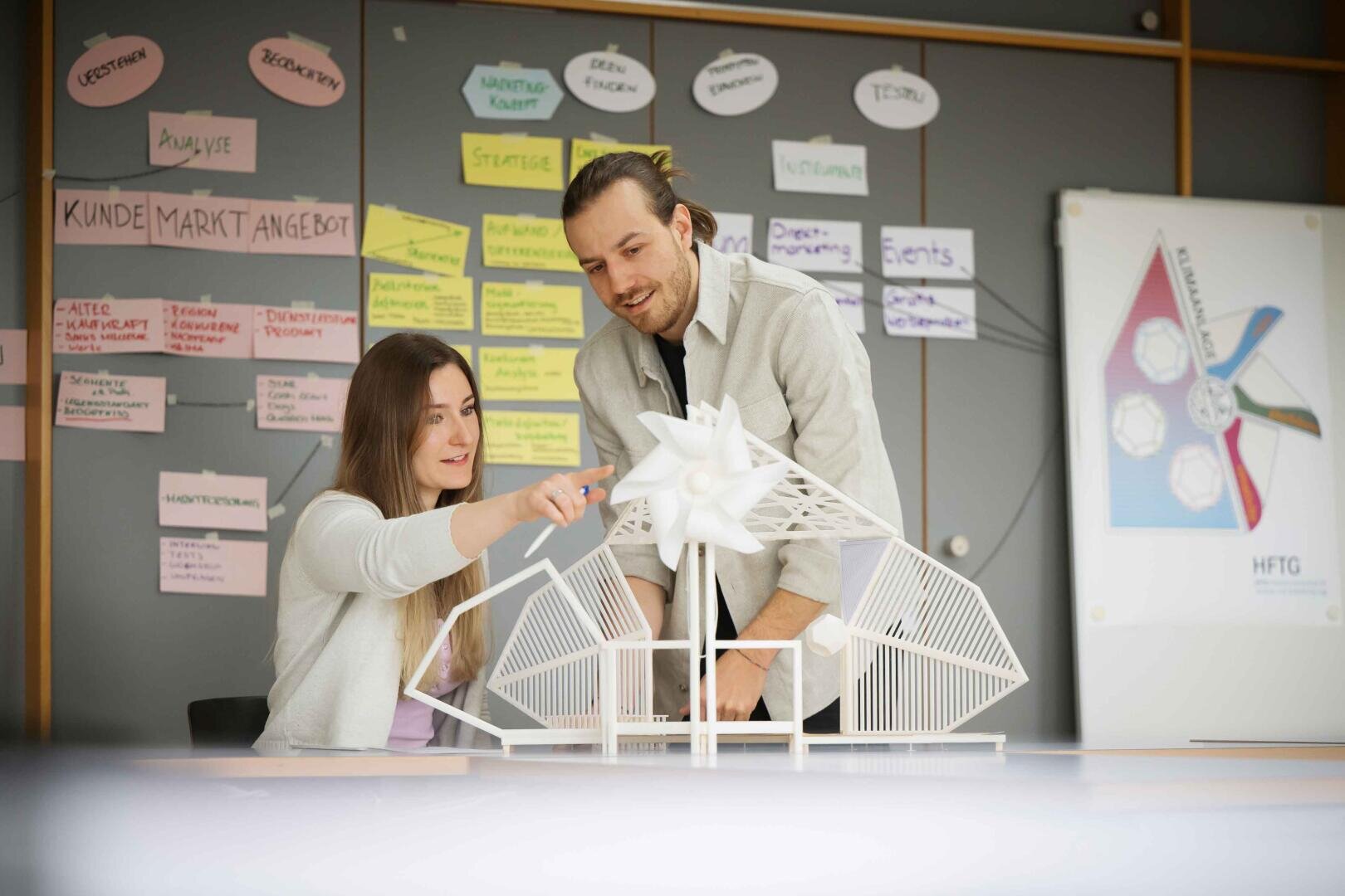 Zwei Personen untersuchen und diskutieren ein detailliertes Architekturmodell auf einem Tisch. An der Wand hinter ihnen sind bunte Notizen und Diagramme zu sehen, die auf eine gemeinsame Planungs- oder Entwurfssitzung hindeuten.