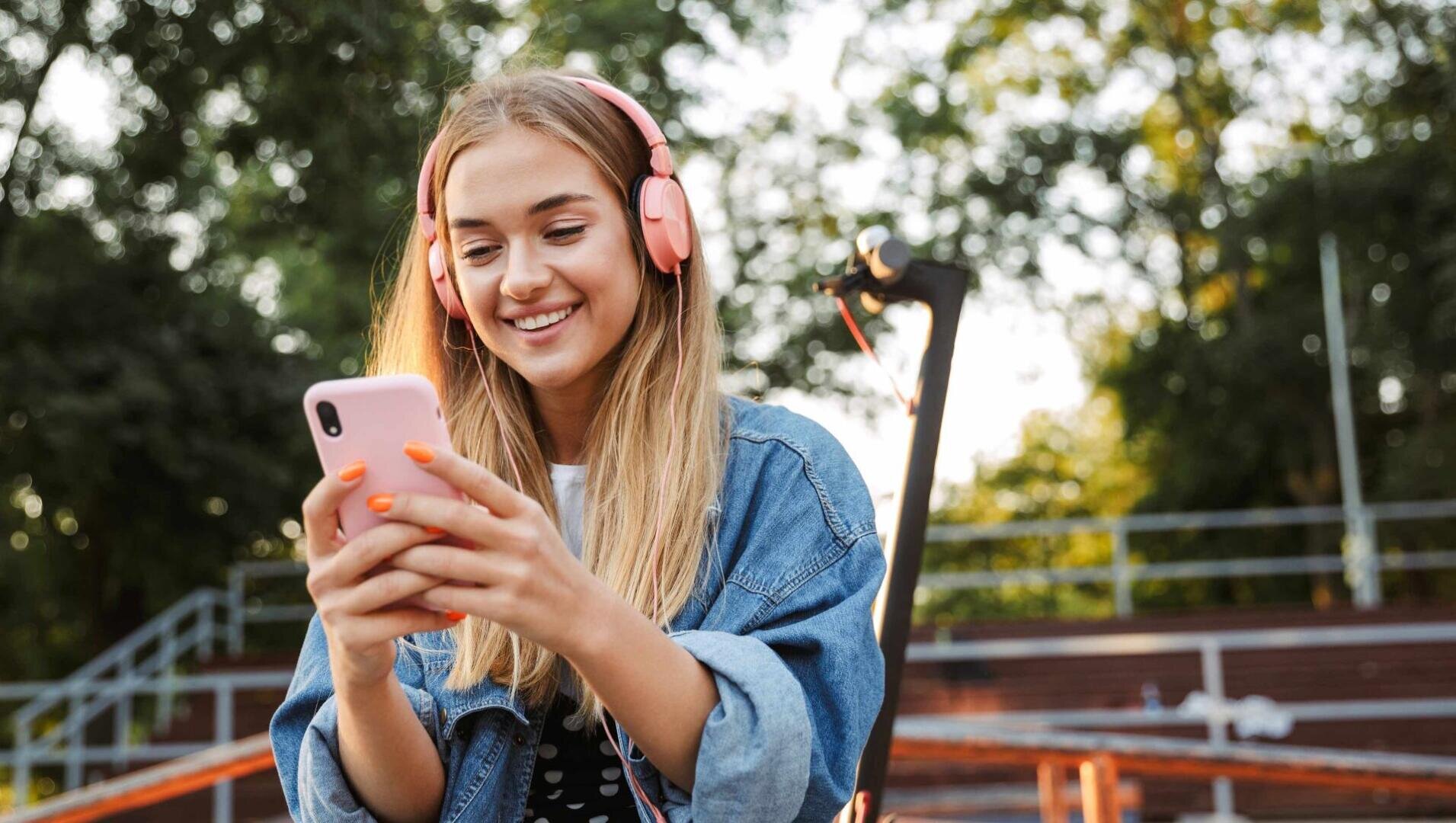 Eine junge Frau mit rosafarbenen Kopfhörern und Jeansjacke lächelt, während sie im Freien auf ihr Smartphone schaut, mit Bäumen und Tribünen im Hintergrund.