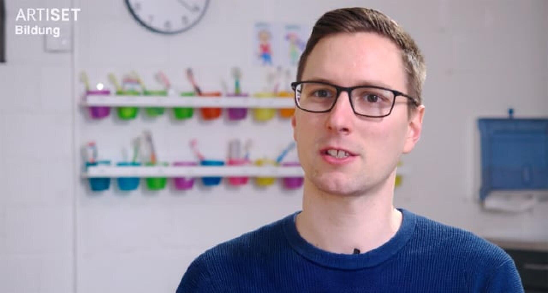 Ein Mann mit Brille und kurzen braunen Haaren, der einen blauen Pullover trägt, sitzt in einem Raum. Hinter ihm stehen bunte Behälter in Regalen, eine Uhr und verschwommene Wanddekorationen. In der oberen linken Ecke ist der Schriftzug „ARTISET Bildung“ zu sehen.