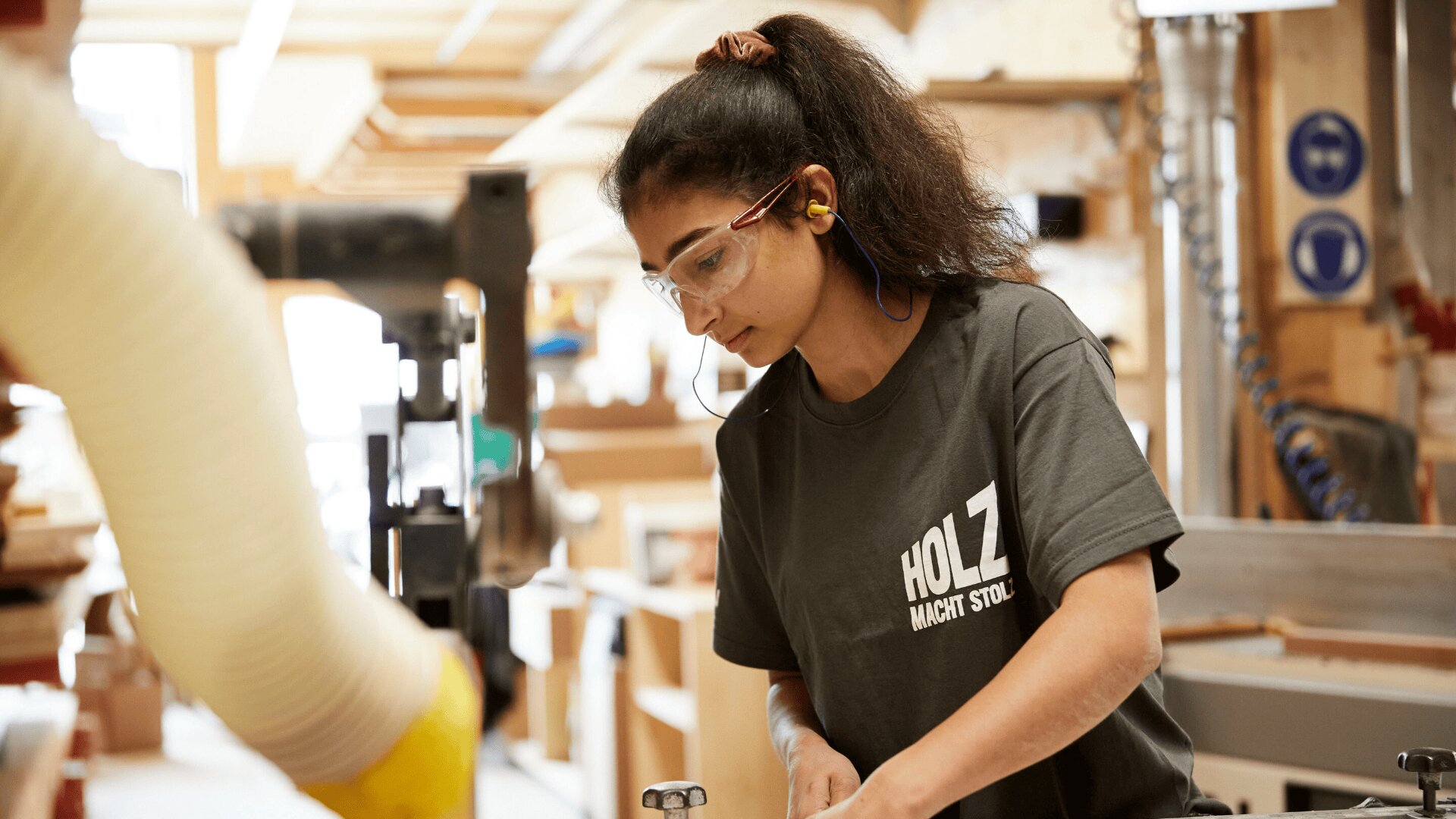 Eine Frau mit Schutzbrille und Ohrstöpseln arbeitet mit Maschinen in einer Holzwerkstatt. Sie ist auf ihre Aufgabe konzentriert und trägt ein graues Hemd mit der Aufschrift HOLZ MACHT STOLZ.