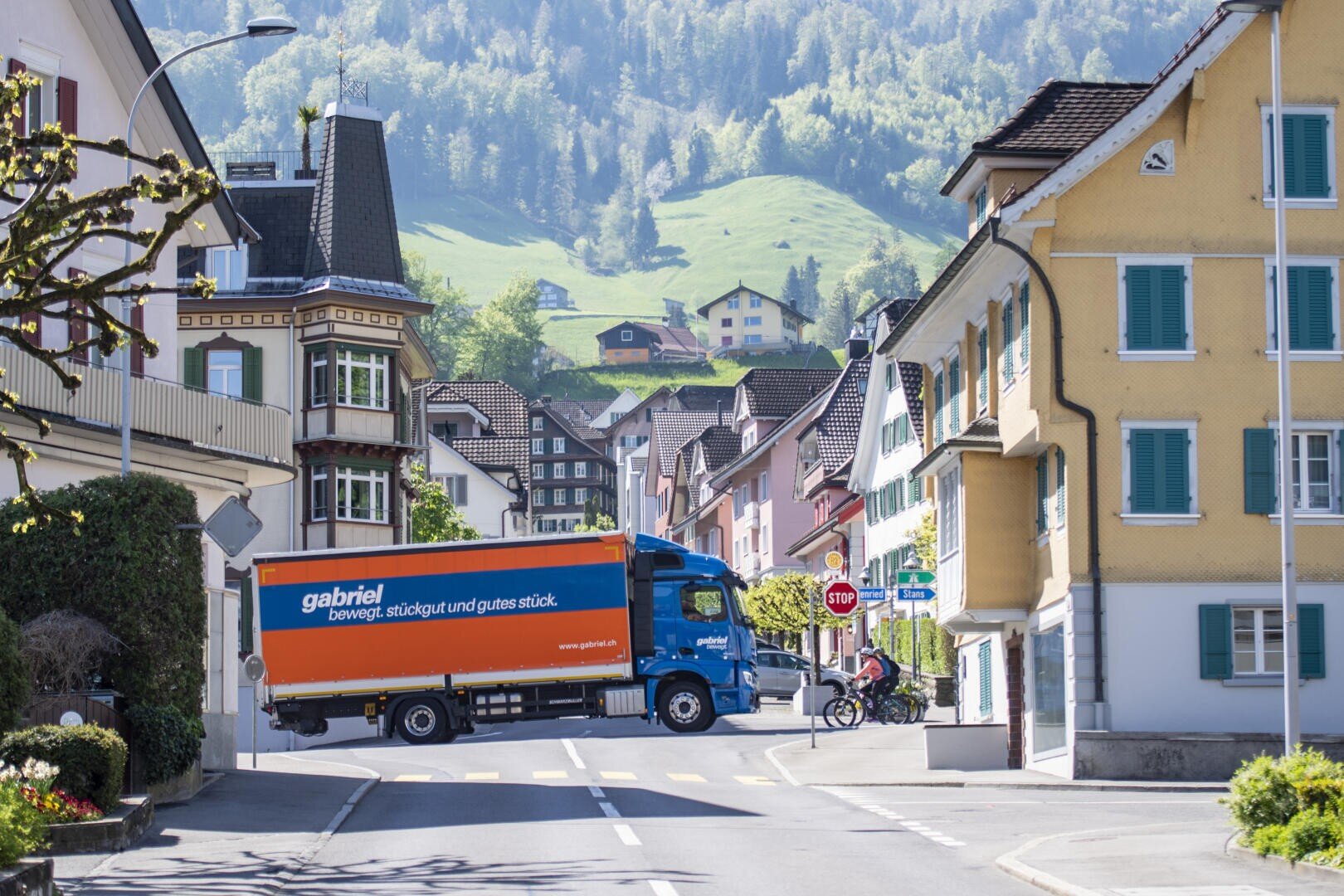 Ein blau-orangefarbener Lieferwagen fährt durch eine ruhige, von bunten Gebäuden gesäumte europäische Dorfstraße, mit grünen Hügeln und verstreuten Häusern im sonnigen Hintergrund.
