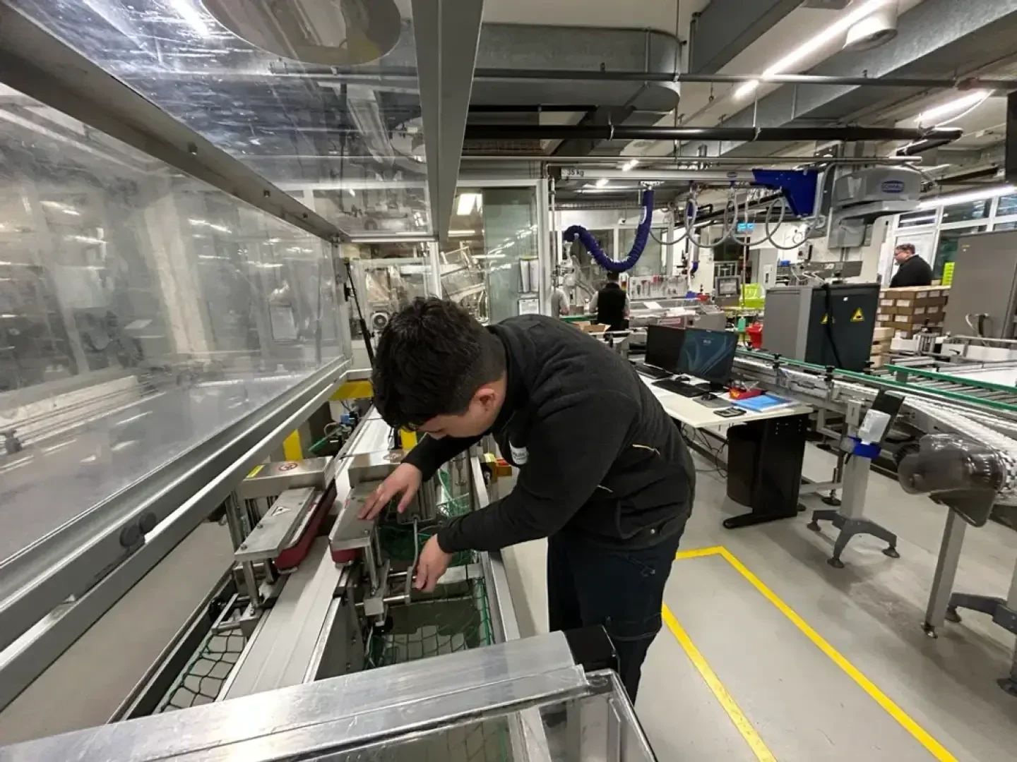 Eine Person arbeitet an einer Maschine in einer geschäftigen Fabrik und justiert Bauteile auf einem Förderband. Der Bereich ist voller Maschinen, Computer und Materialvorräte; im Hintergrund ist ein weiterer Arbeiter zu sehen.
