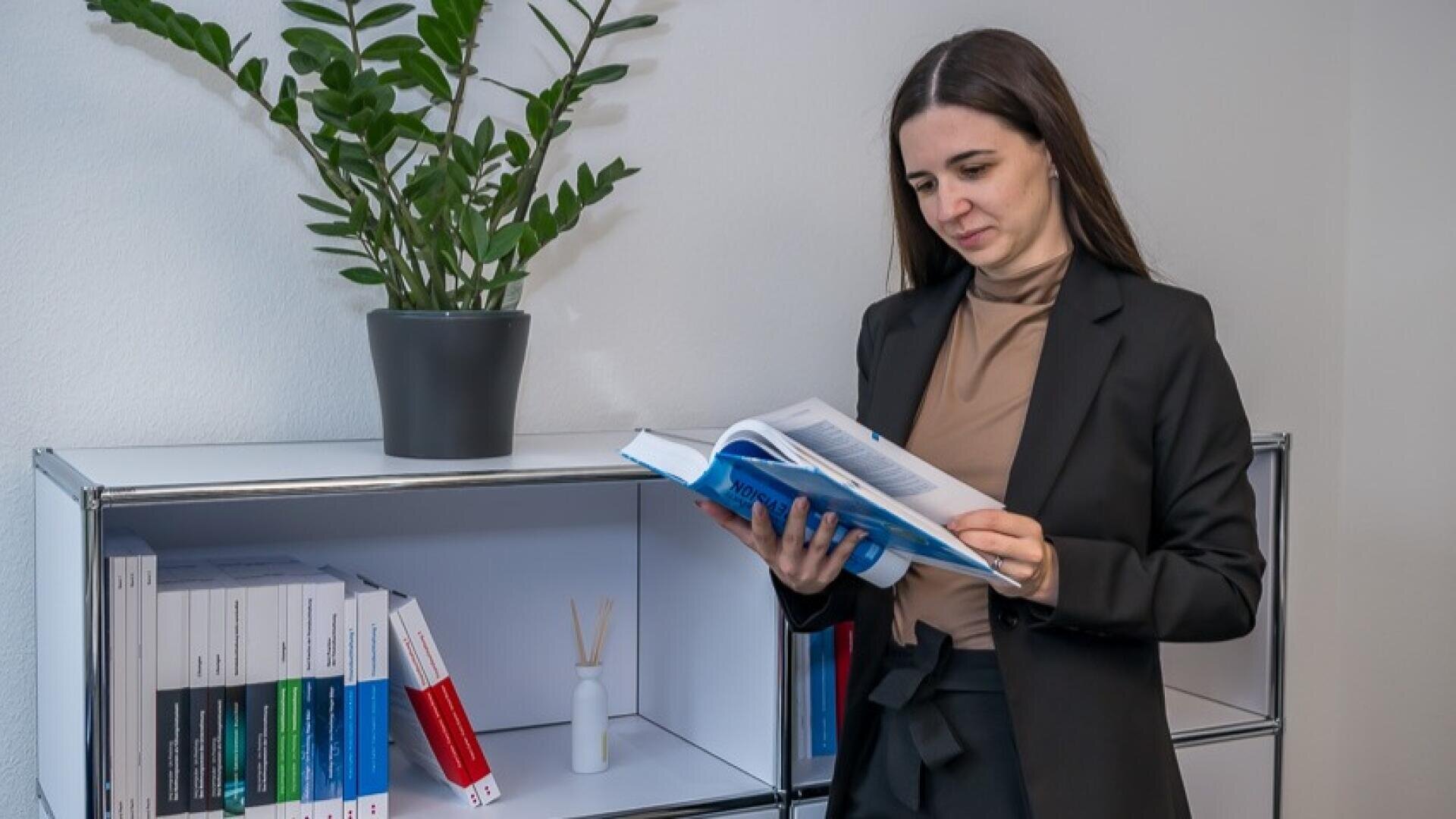 Eine Frau in einem schwarzen Blazer, die vor einem Regal mit Büchern und einer Topfpflanze aus einem aufgeschlagenen Buch liest.