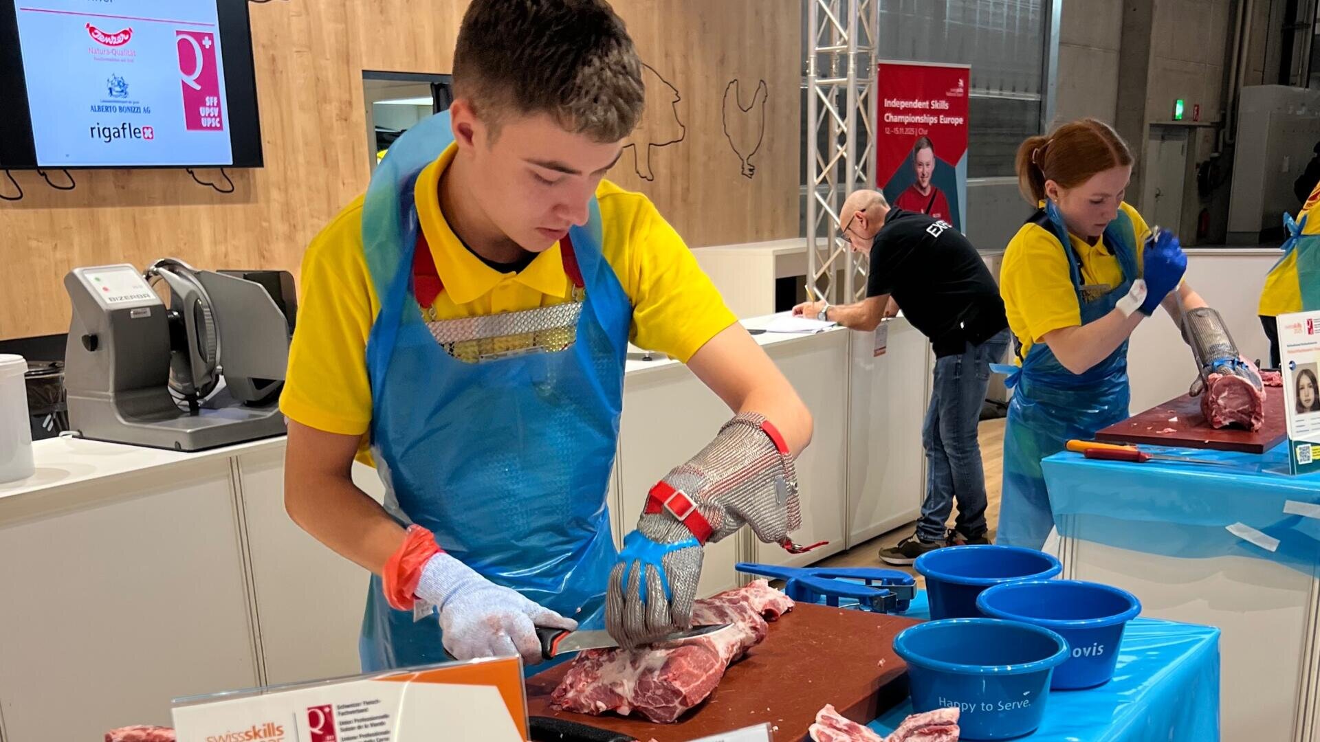 Zwei junge Menschen in Schutzkleidung und gelben Hemden schneiden an getrennten Arbeitsstationen im Rahmen eines Wettbewerbs Fleisch, während im Hintergrund die Juroren und die Ausrüstung zu sehen sind.