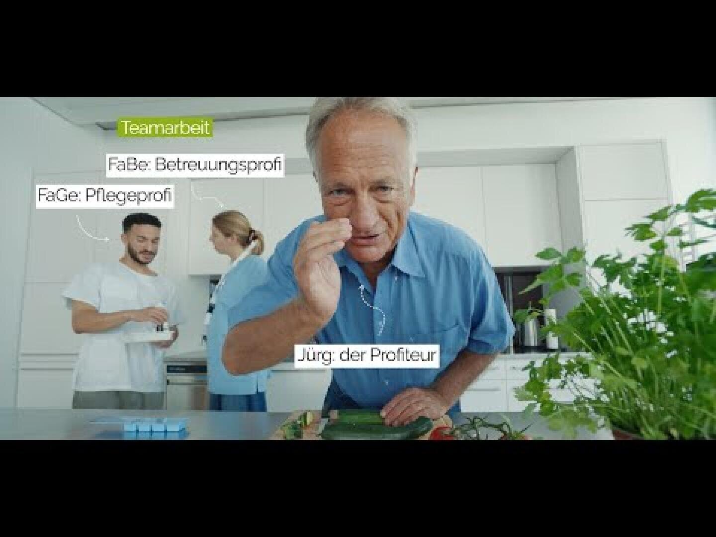 Drei Personen in einer Küche: Ein Mann im Vordergrund winkt in die Kamera, während zwei andere in weißen Uniformen Essen zubereiten. Deutsche Beschriftungen beschreiben ihre Rollen: „Teamarbeit“, „Pflegeprofi“, „Betreuungsprofi“ und „Jürg der Profiteur“.