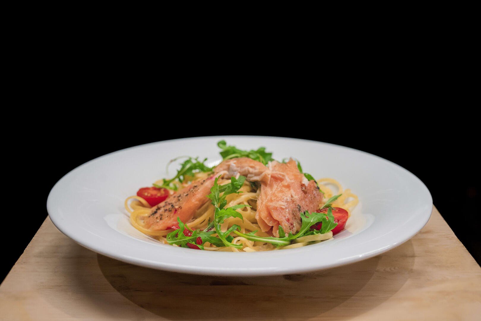 Ein weißer Teller mit Nudeln, die mit Lachsscheiben, Rucola und Kirschtomaten belegt sind, steht auf einer Holzplatte vor einem schwarzen Hintergrund.