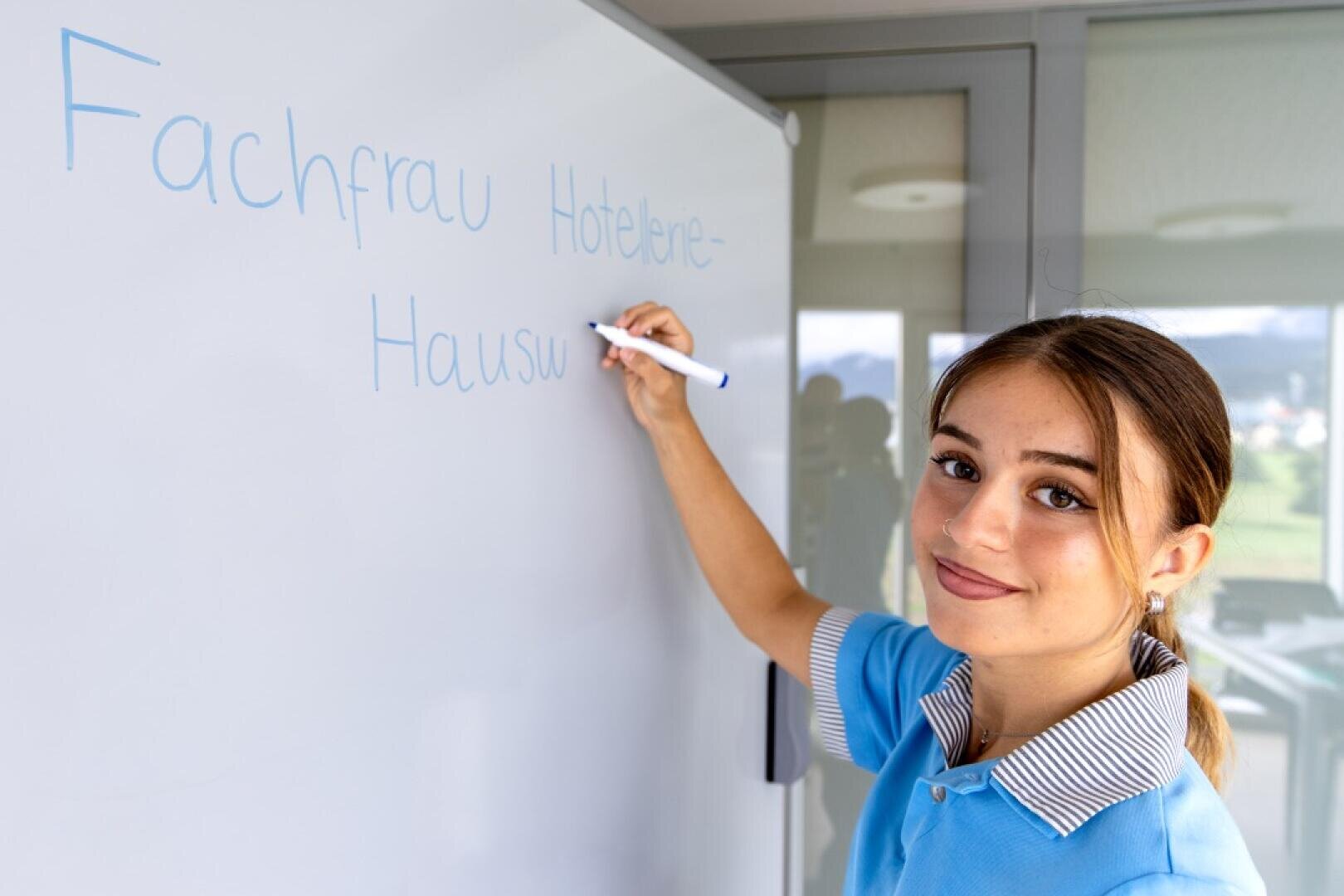 Eine junge Frau in einer blauen Uniform schreibt mit einem blauen Marker Fachfrau Hotelerie-Hausw... auf eine Tafel und lächelt in die Kamera, in einem hellen Innenraum mit einem Fenster im Hintergrund.
