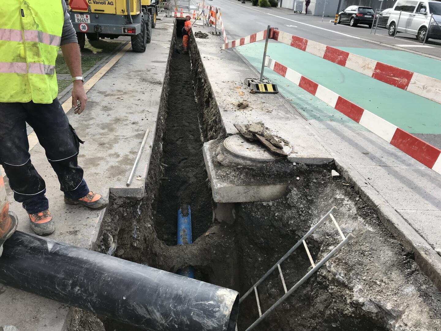 Arbeiter verlegen oder reparieren unterirdische Leitungen in einem langen, schmalen Graben neben einer Straße. Es sind Verkehrsabsperrungen und Baumaschinen vorhanden, und eine Leiter führt in den Graben hinunter.