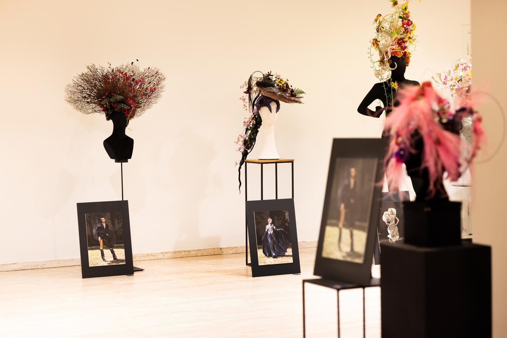 Eine Galerie mit Schaufensterpuppen, die kunstvolle Blumenkopfbedeckungen tragen, und gerahmten Modefotografien, die auf dem Boden vor einer schlichten, hellen Wand stehen.