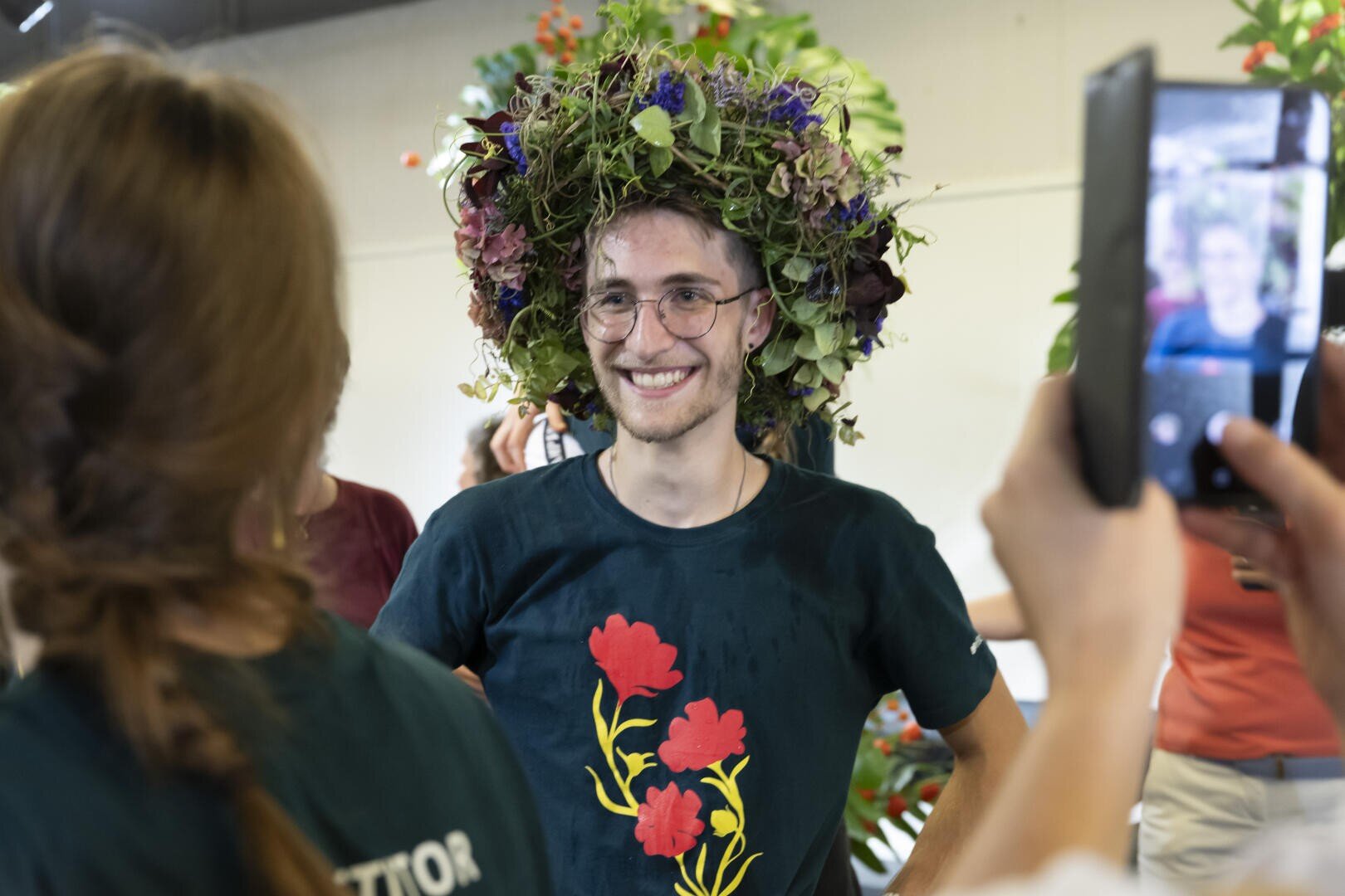 Eine lächelnde Person mit Brille und einer großen, bunten Blumenkrone steht in einem Haus. Sie trägt ein dunkles Hemd mit roten Blumen. Andere um sie herum machen Fotos, und die Stimmung ist fröhlich.