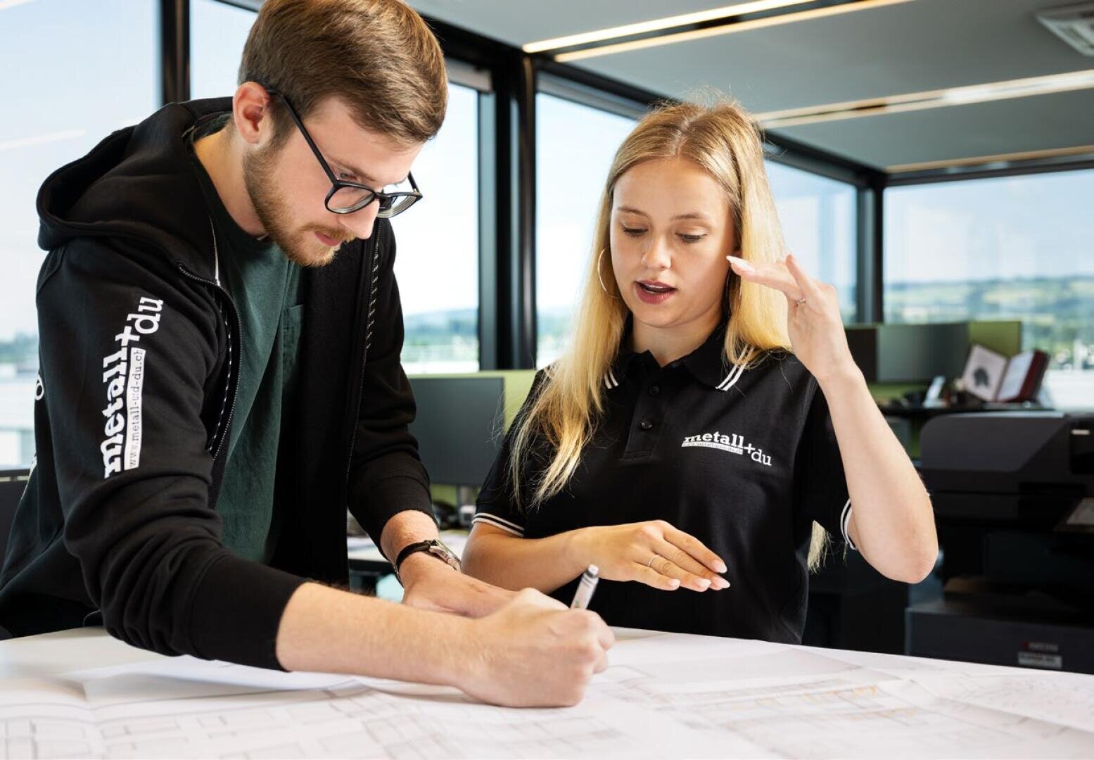 Zwei junge Berufstätige begutachten Architekturpläne auf einem Schreibtisch in einem modernen Büro. Der Mann schreibt Notizen, während die Frau gestikuliert, als würde sie etwas erklären. Große Fenster sorgen für natürliches Licht im Hintergrund.