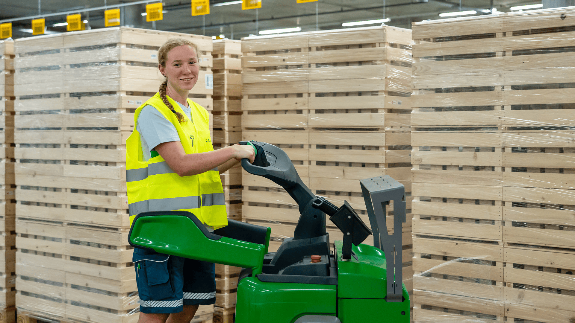 Eine Person in einer gelben Sicherheitsweste und Shorts bedient eine grüne Maschine in einem Lagerhaus, umgeben von Stapeln aus mit Plastikfolie umwickelten Holzpaletten.
