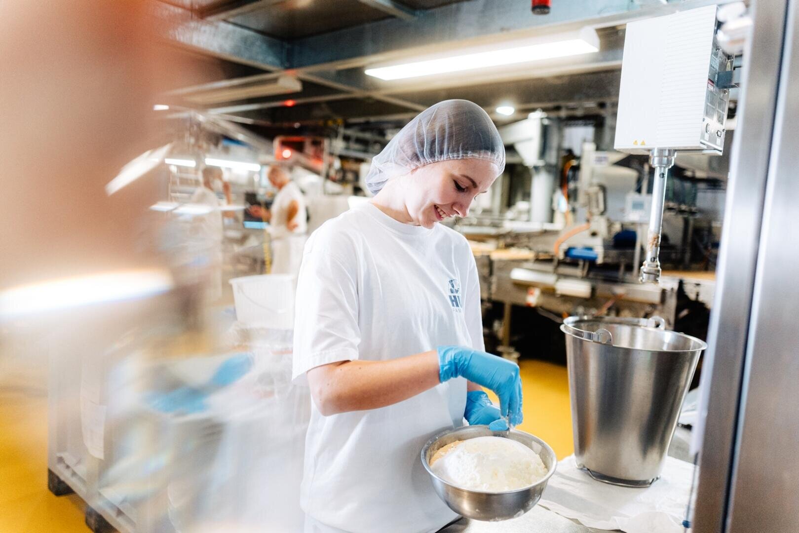 Eine Person, die ein Haarnetz, Handschuhe und ein weißes T-Shirt trägt, mischt Zutaten in einer Metallschüssel in einer Großküche oder Lebensmittelproduktionsanlage. Im Hintergrund sind Industrieanlagen und ein weiterer Arbeiter zu sehen.