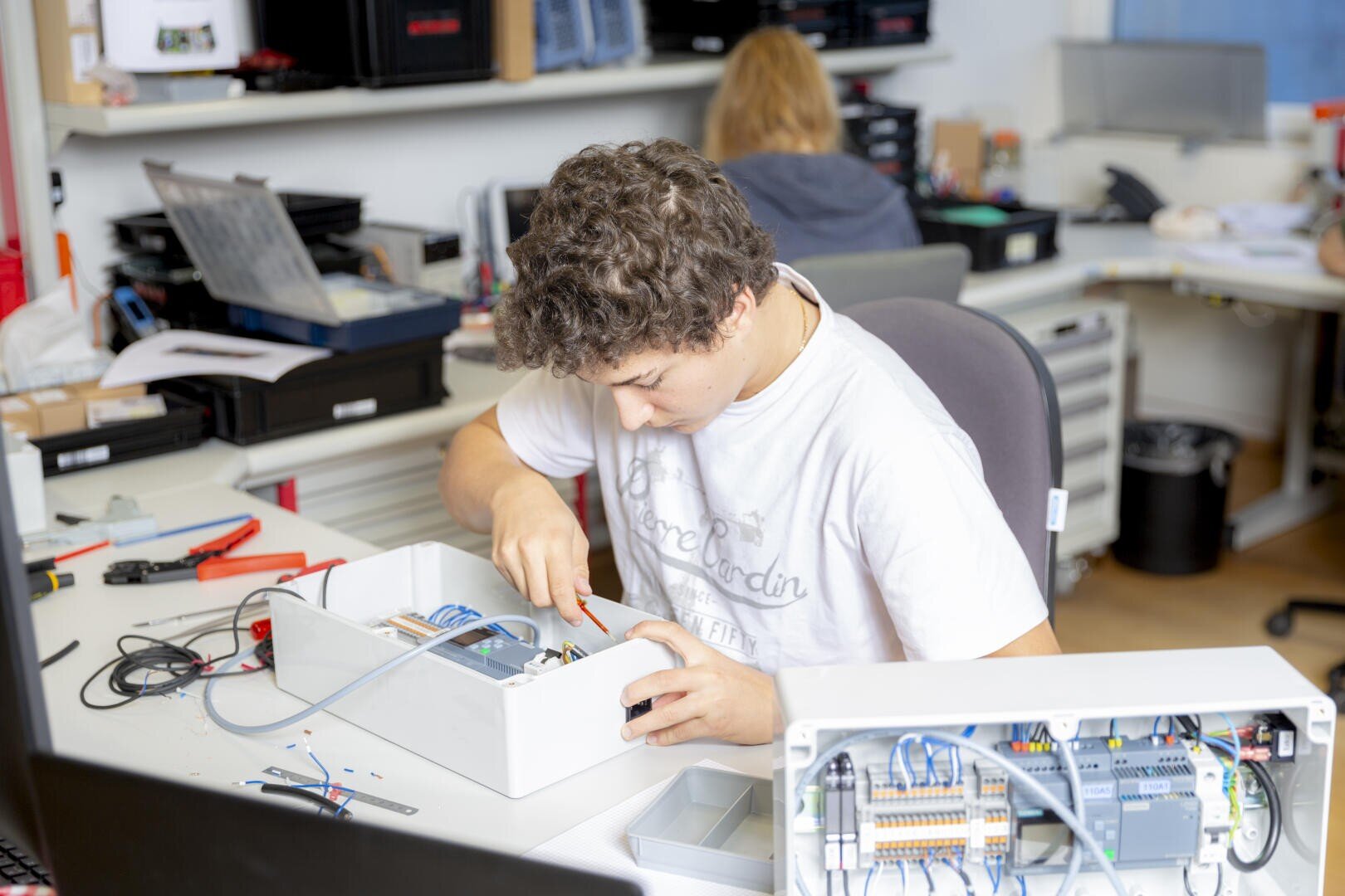 Eine junge Person mit lockigem Haar baut an einem Schreibtisch mit Werkzeugen elektronische Komponenten in einem weißen Gehäuse zusammen. Eine andere Person arbeitet an einem Computer im Hintergrund in einem hellen, organisierten Arbeitsbereich.